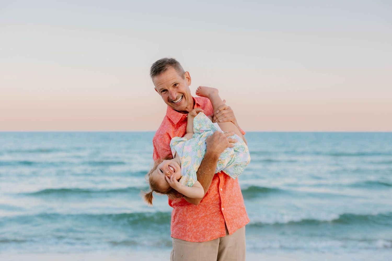 Man in coral shirt joyfully lifting baby on a sunny beach with calm ocean waves in the background.
