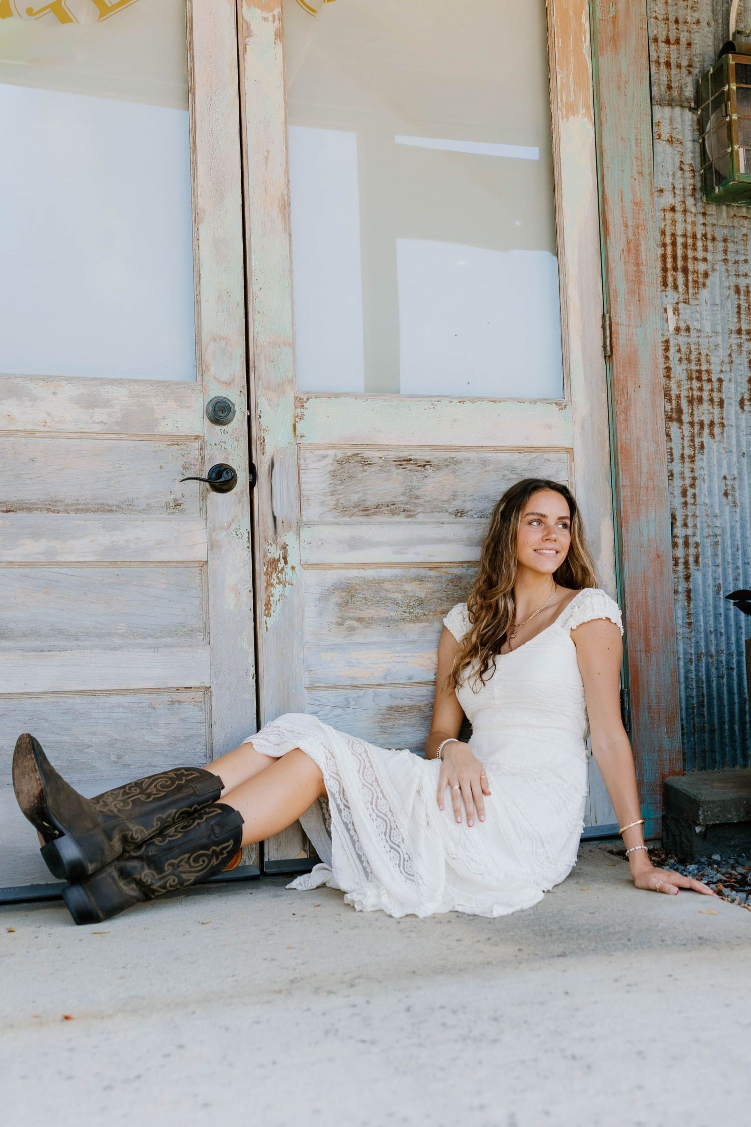 Woman in white lace dress and cowboy boots sitting against rustic wooden doors, smiling outdoors.