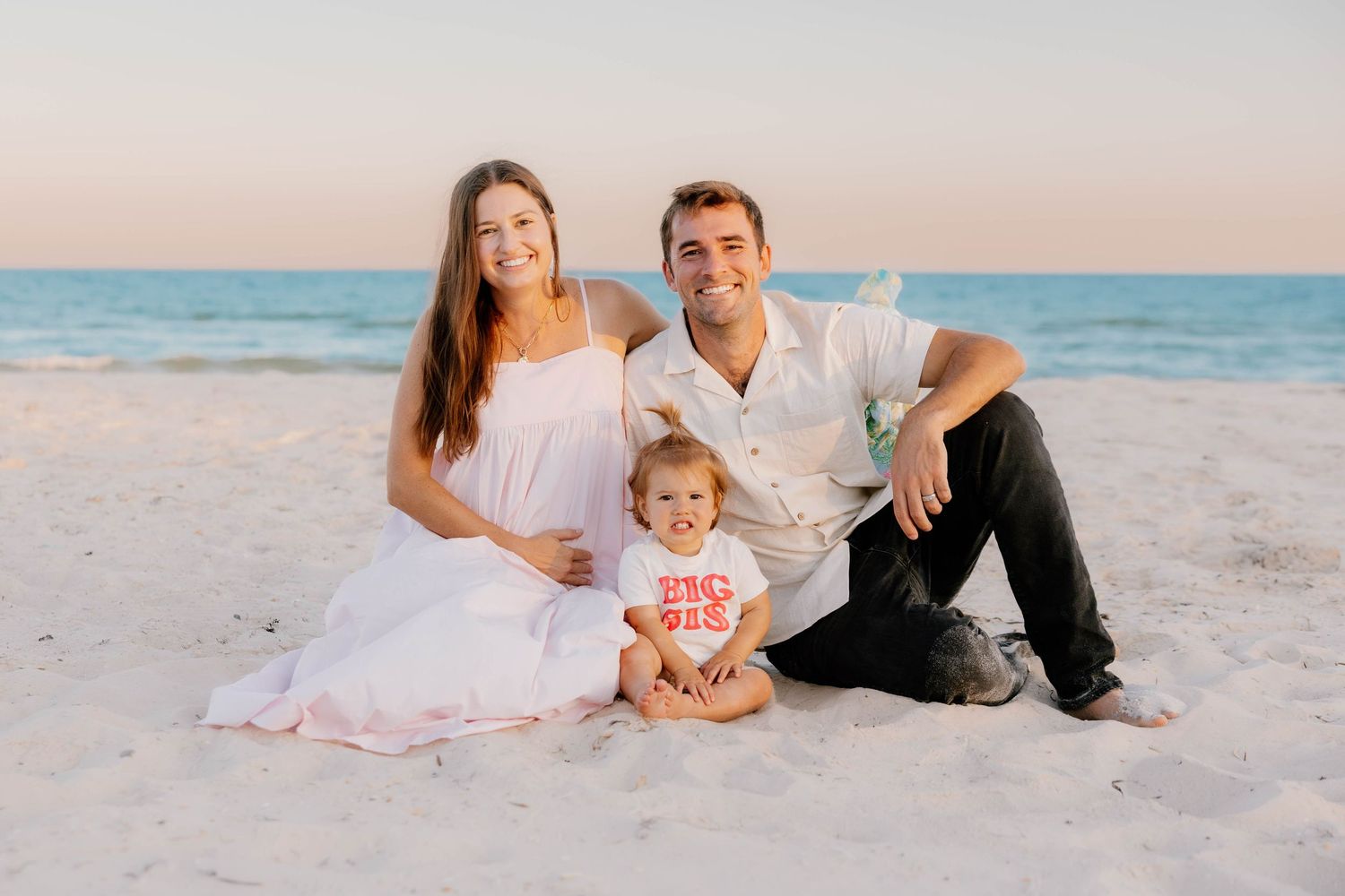 Family of three sitting on sandy beach at sunset, parents smiling with toddler in red shirt, ocean backdrop.