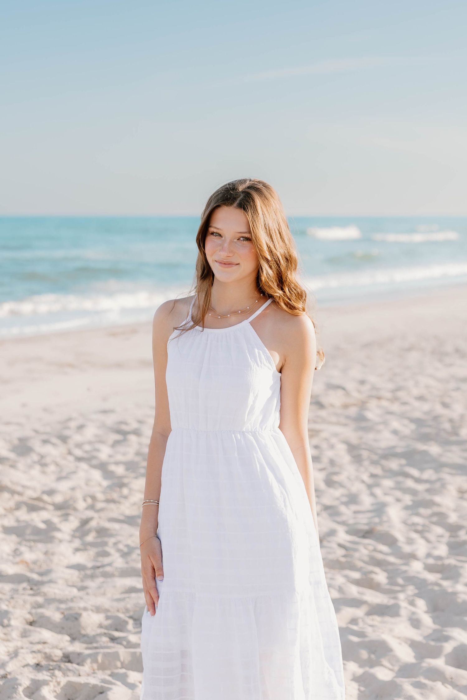 Young woman in white sundress standing on sandy beach with ocean waves and blue sky in background.