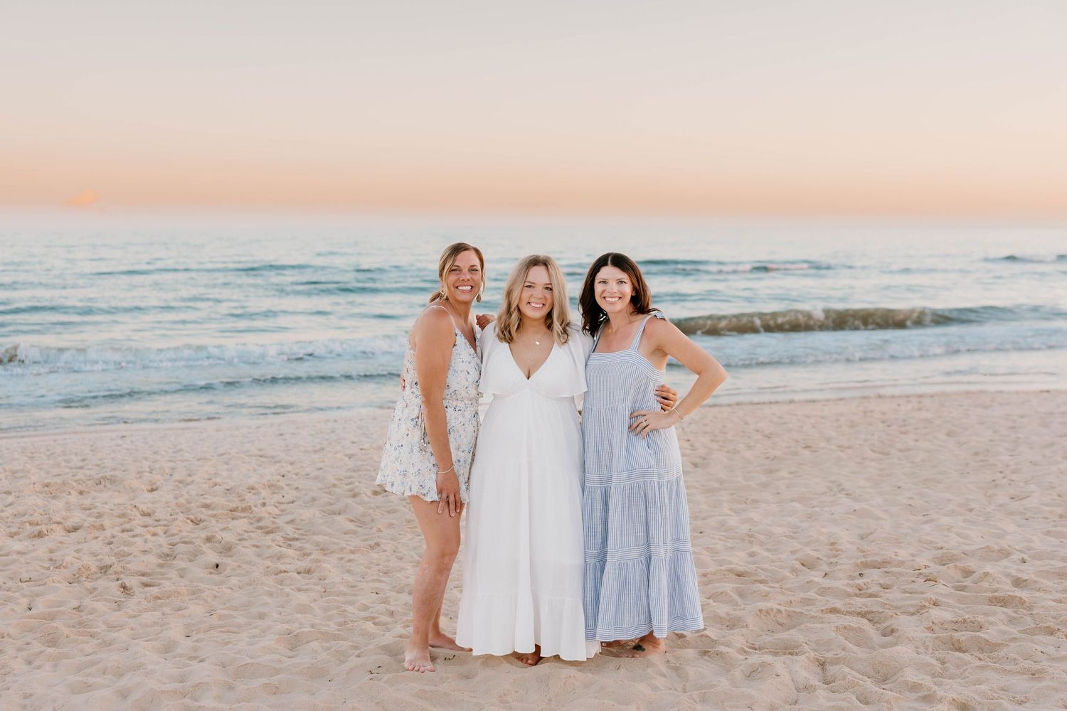 Three women smiling together on a sandy beach at sunset wearing white and blue dresses.