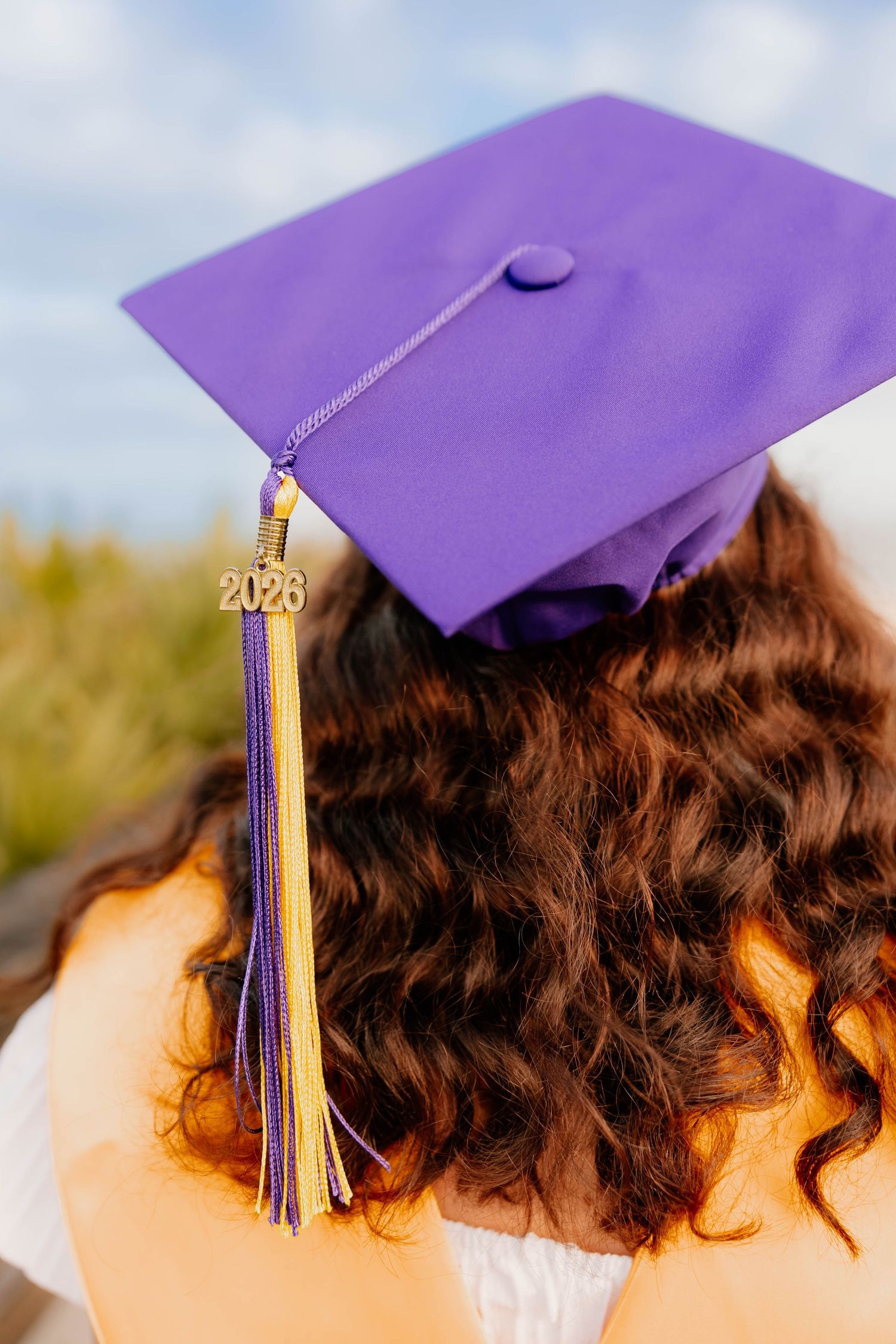 Graduate wearing purple mortarboard cap with gold 2026 tassel and curly brown hair outdoors.