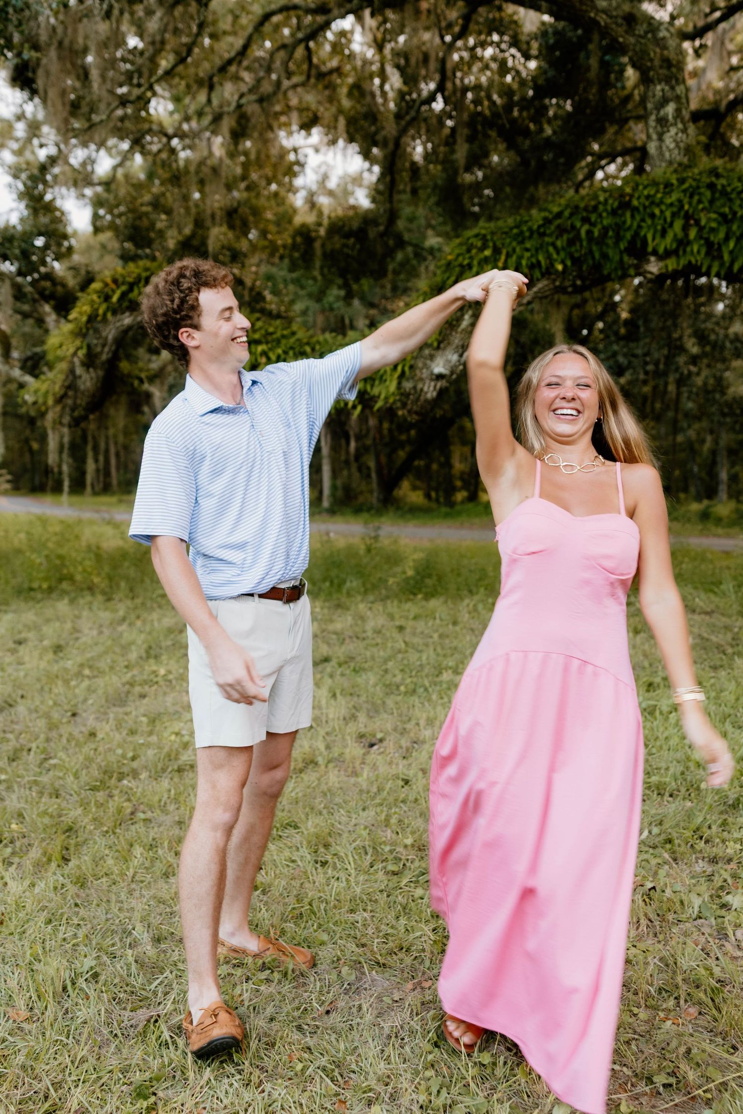 A joyful couple dancing outdoors, man in blue shirt spinning woman in pink maxi dress among lush green trees.