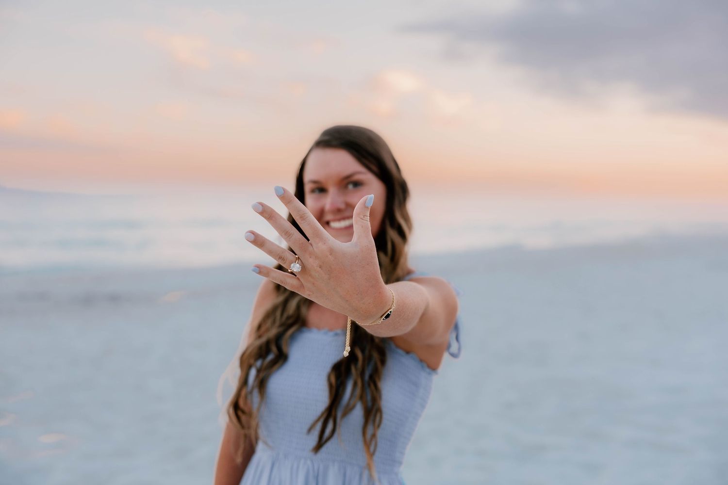 Woman in white dress showing off engagement ring on beach at sunset with ocean backdrop.
