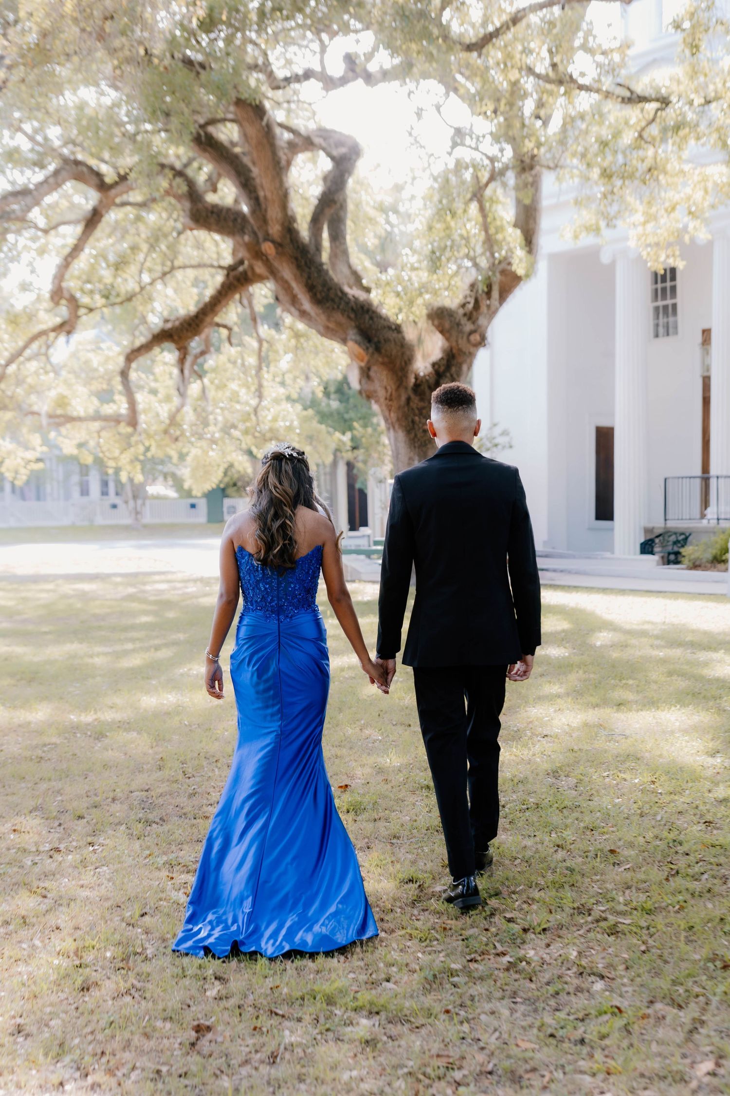 Couple walking hand in hand, woman in blue mermaid gown and man in black tuxedo, beneath a large oak tree.