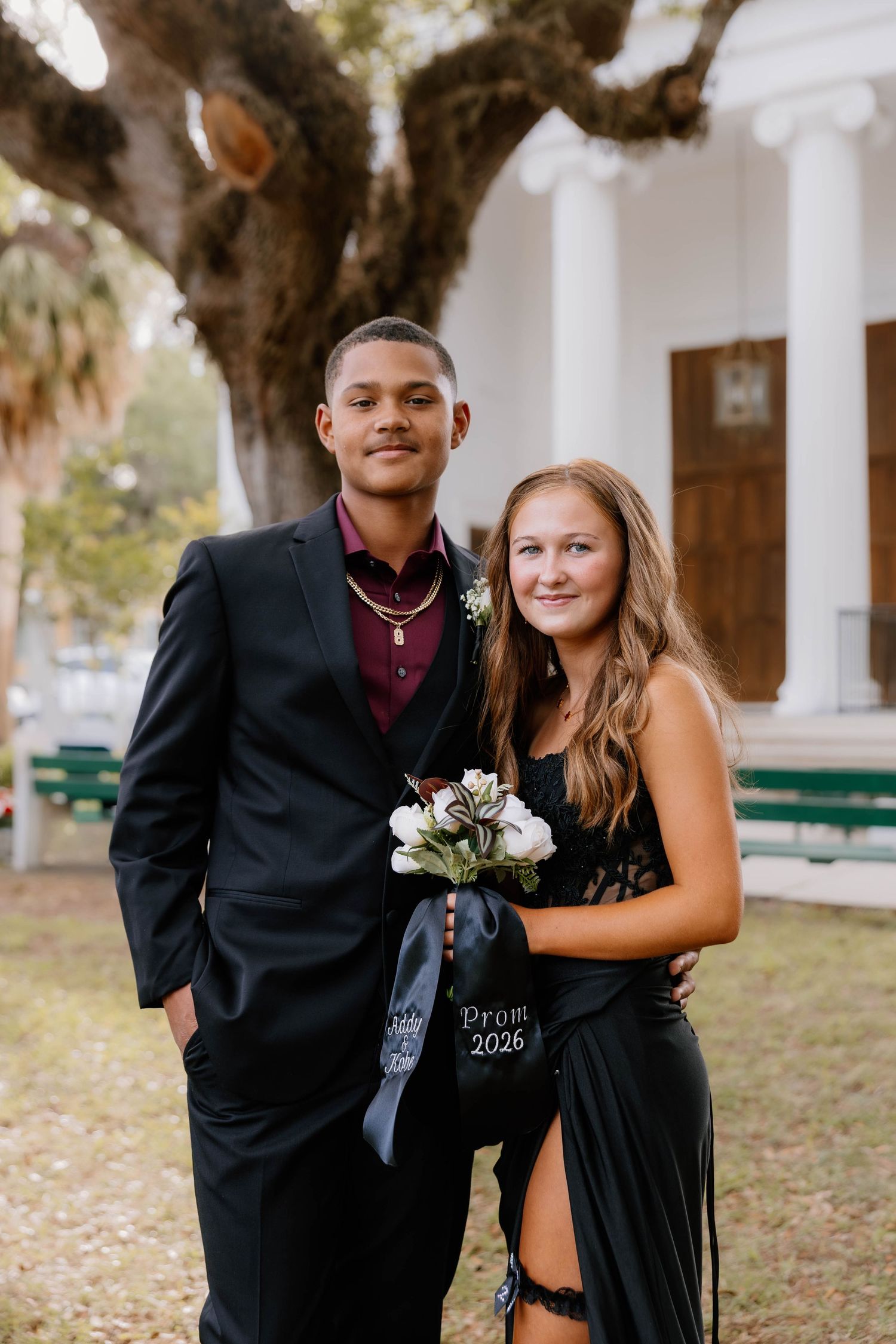 Two prom-dressed teens pose outdoors, girl in black gown holding bouquet, boy in suit with boutonniere, Prom 2026 sash.