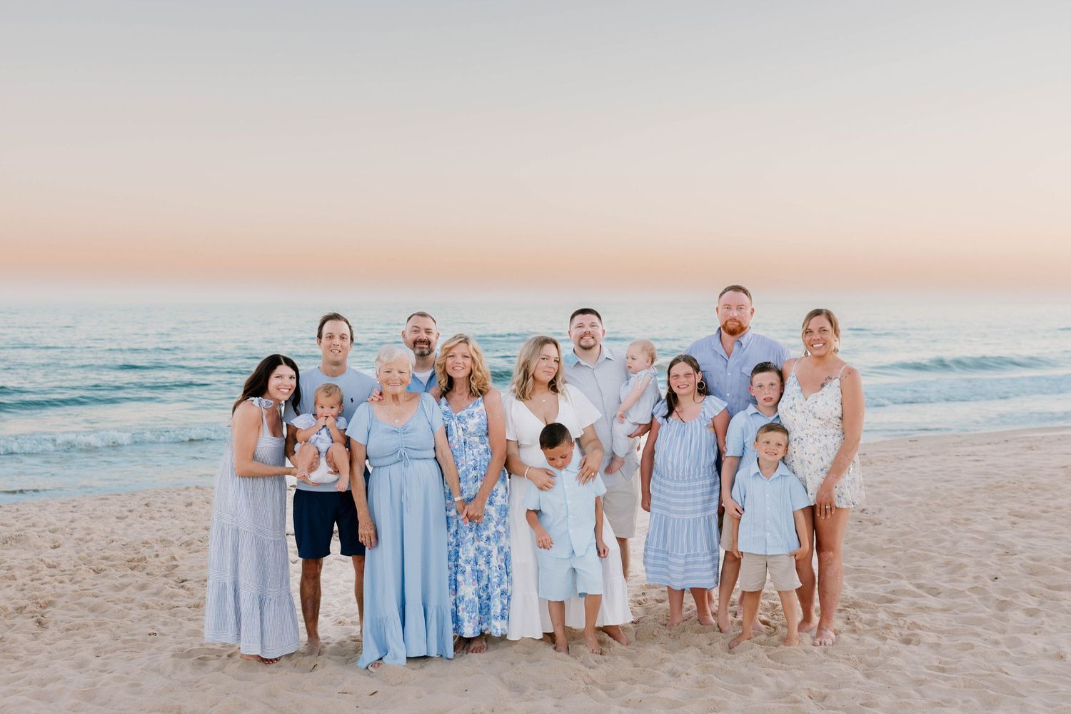 Large family posing together on a sandy beach at sunset, wearing coordinated blue and white outfits.