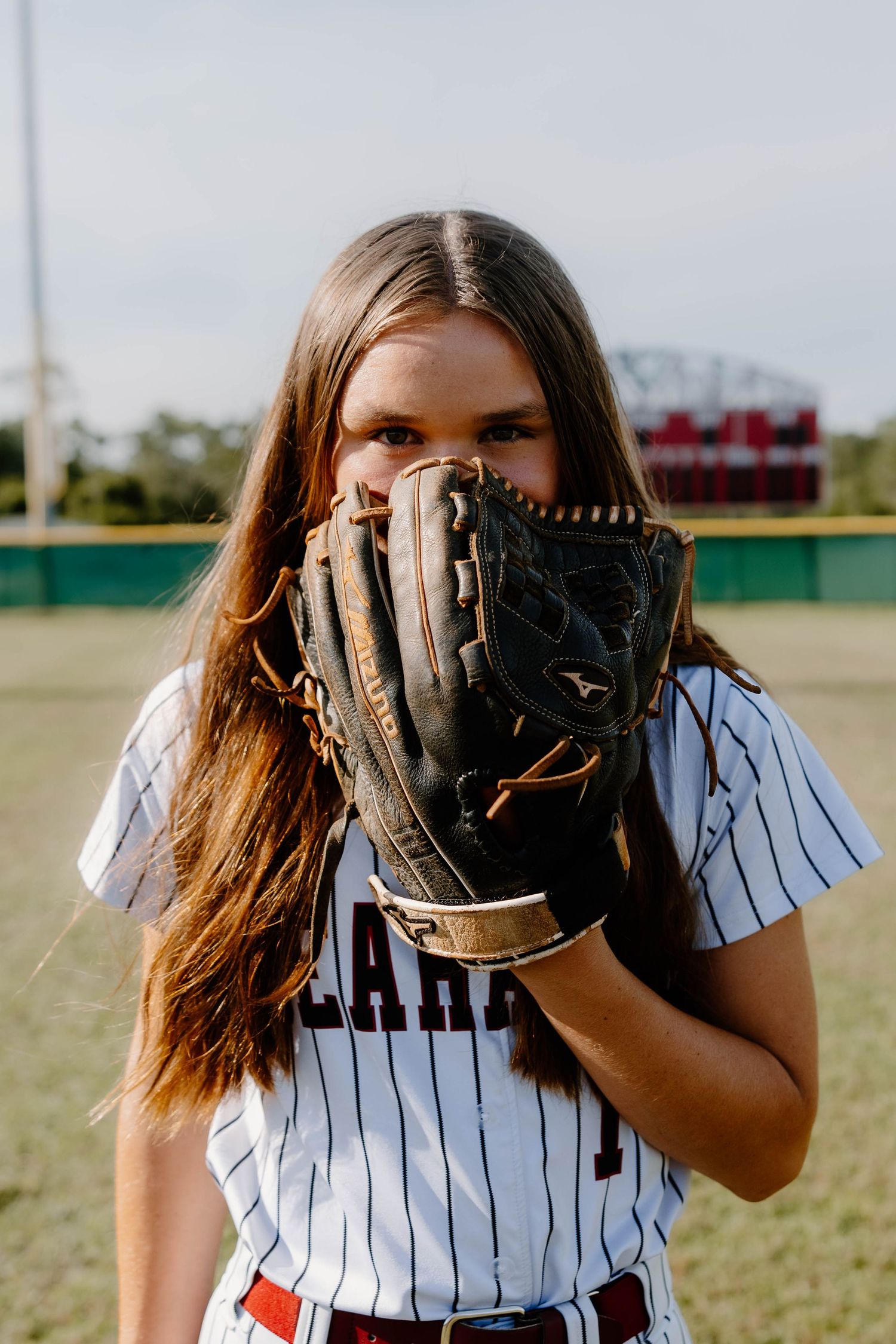 Young softball player in pinstripe uniform holds glove up to face on baseball field.