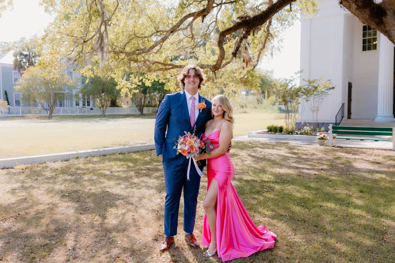Couple posing outdoors in formal attire, man in navy suit and woman in pink gown holding floral bouquet.