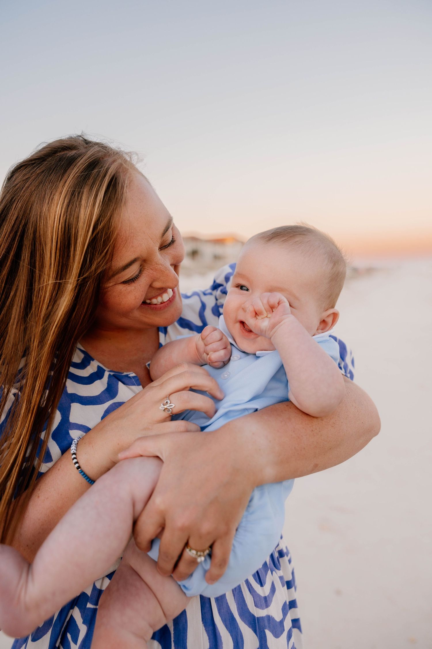 Smiling mother holding happy baby on beach at sunset, both wearing blue and white outfits.
