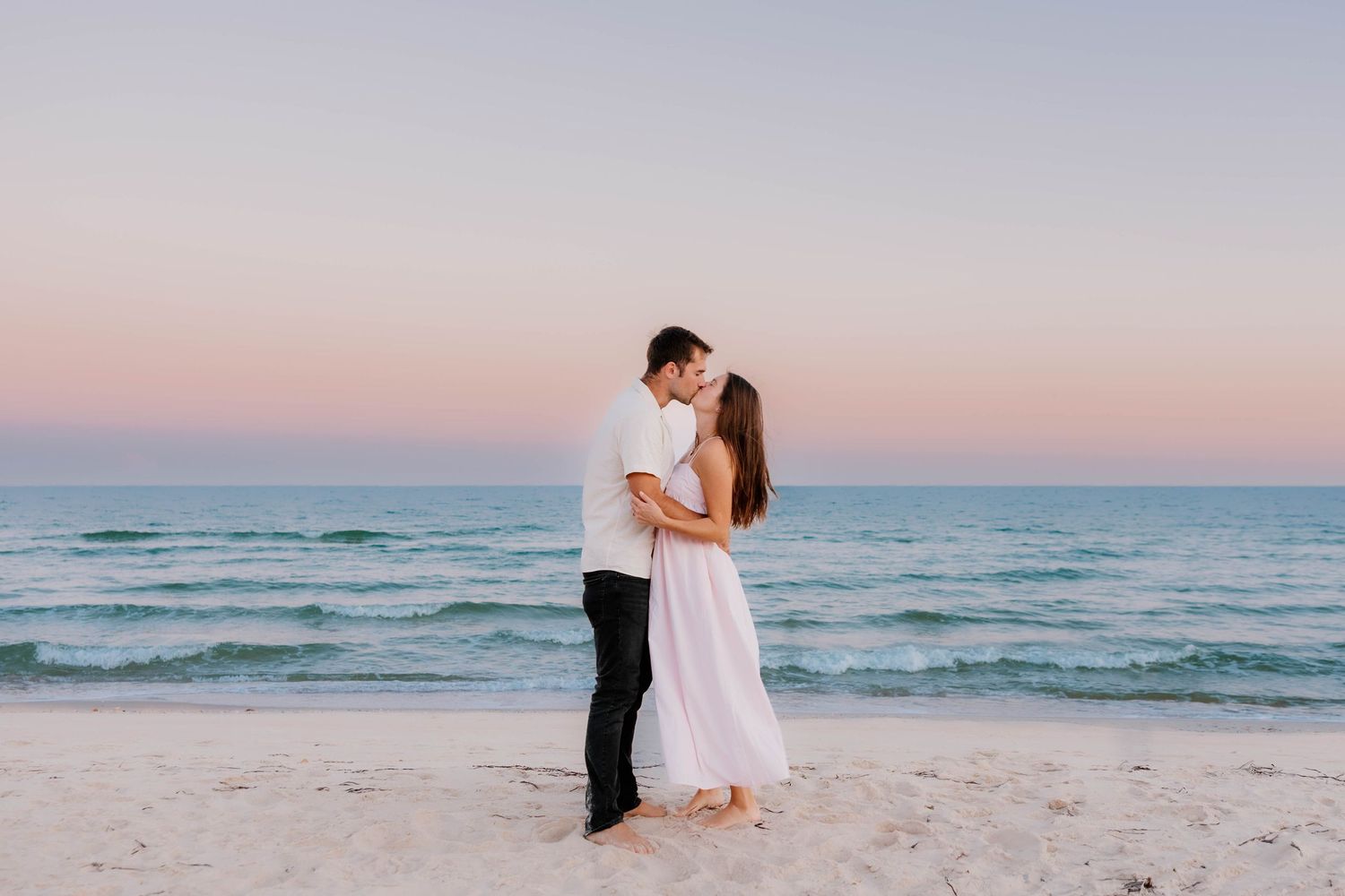 Couple embracing on a sandy beach at sunset with turquoise ocean waves and pink sky backdrop.