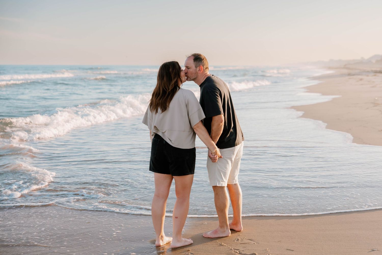 Couple kissing on a sandy beach at sunset, holding hands with ocean waves in the background.