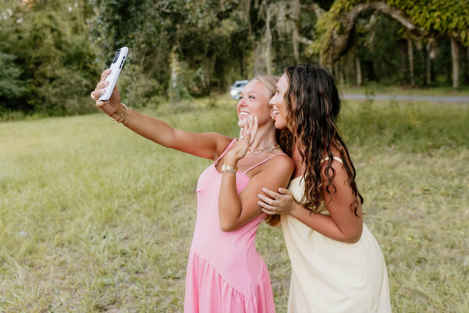 Two women in pink and yellow dresses take a selfie outdoors in a green field surrounded by trees.