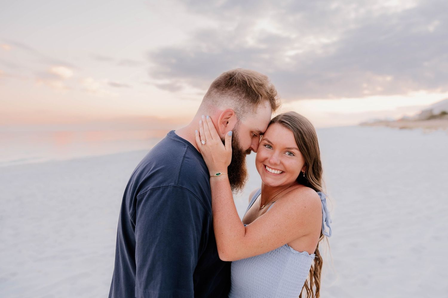 Couple sharing a romantic beach moment at sunset, woman smiling as man nuzzles her forehead on sandy shore.