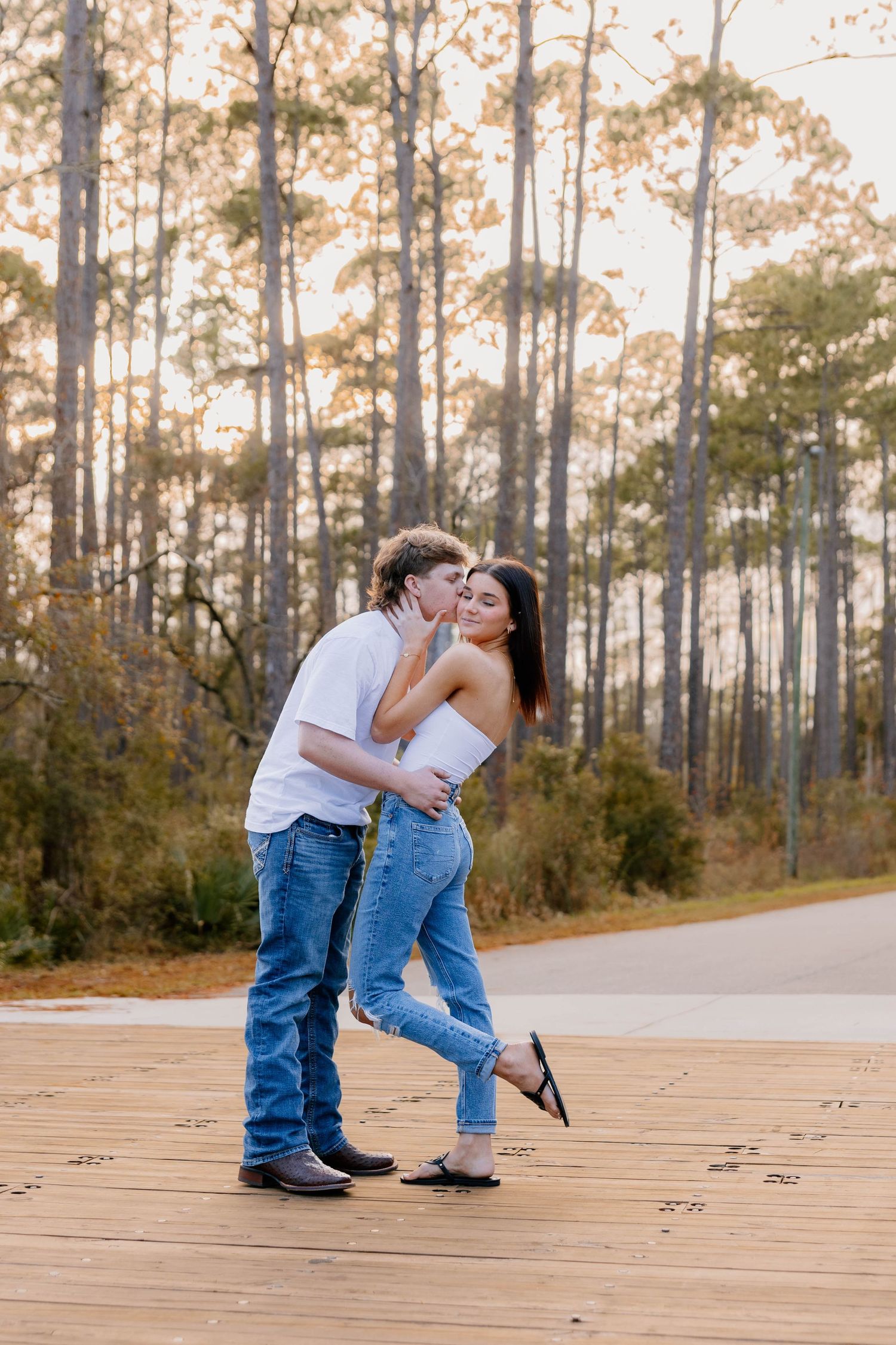 A young couple sharing a kiss on a wooden deck surrounded by tall pine trees at golden hour.