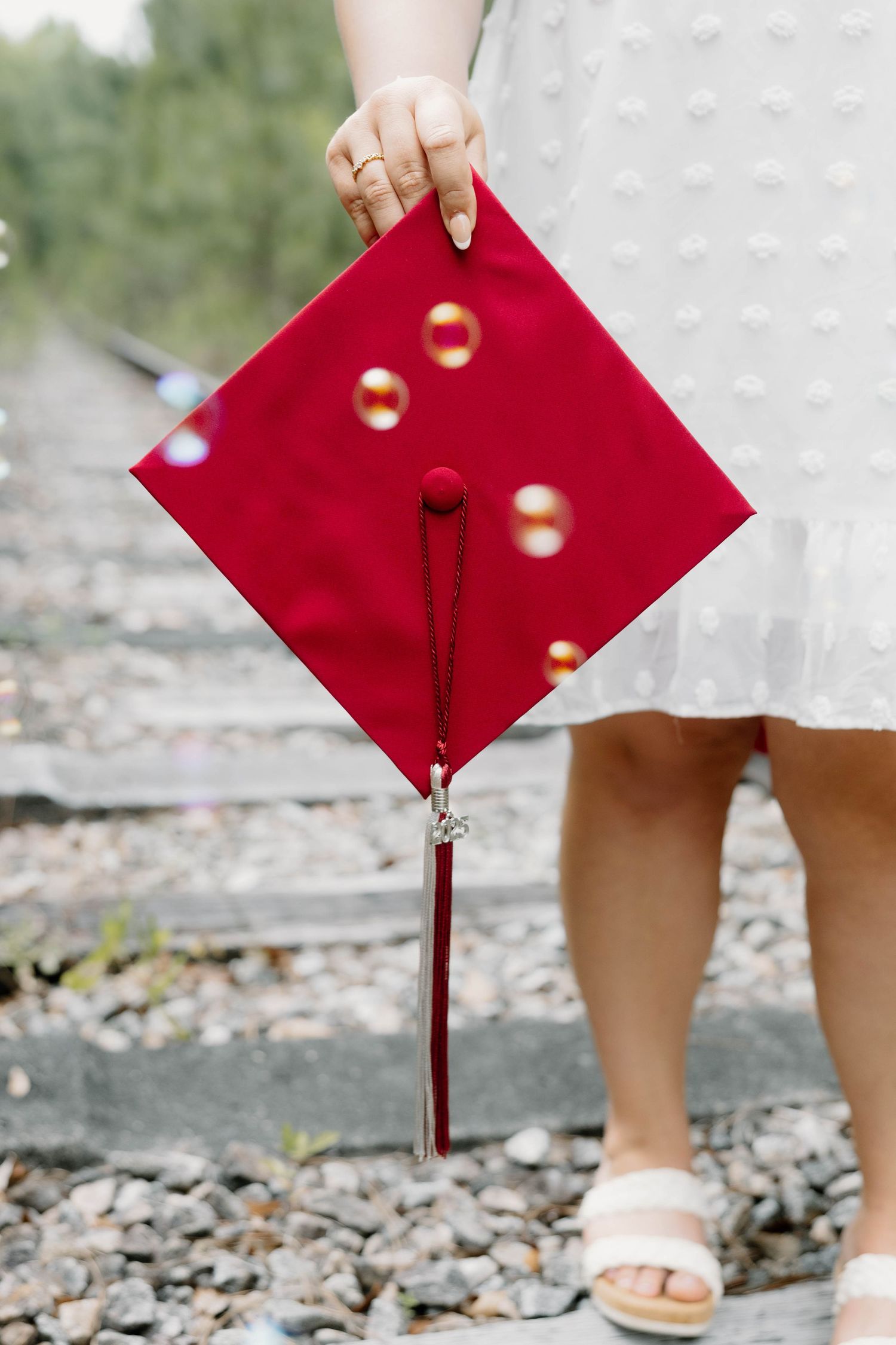 Person holding a red graduation cap with tassel and bubbles, standing on gravel railroad tracks in a white dress.