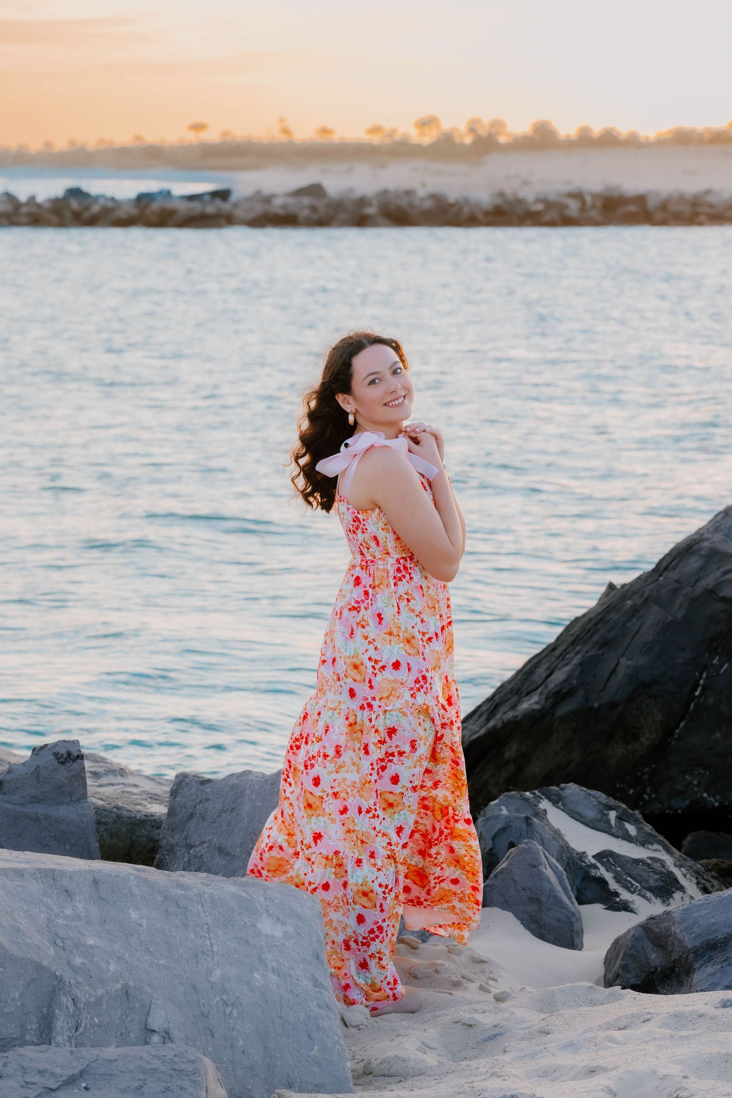 Woman in floral maxi dress smiling on rocky beach at golden hour sunset near calm ocean waters.