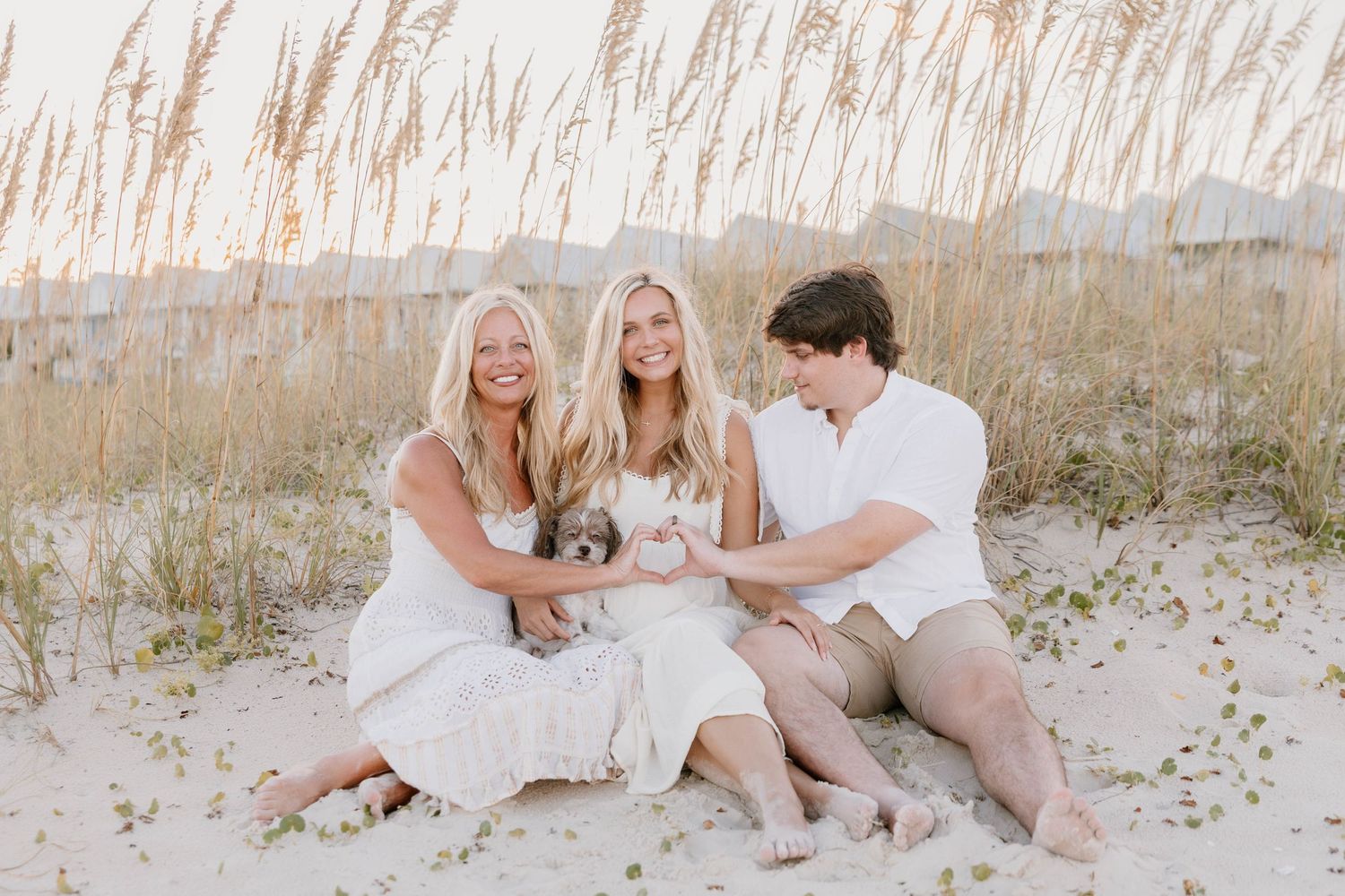 A family wearing white and beige outfits poses together on a sandy beach with tall grass in the background.
