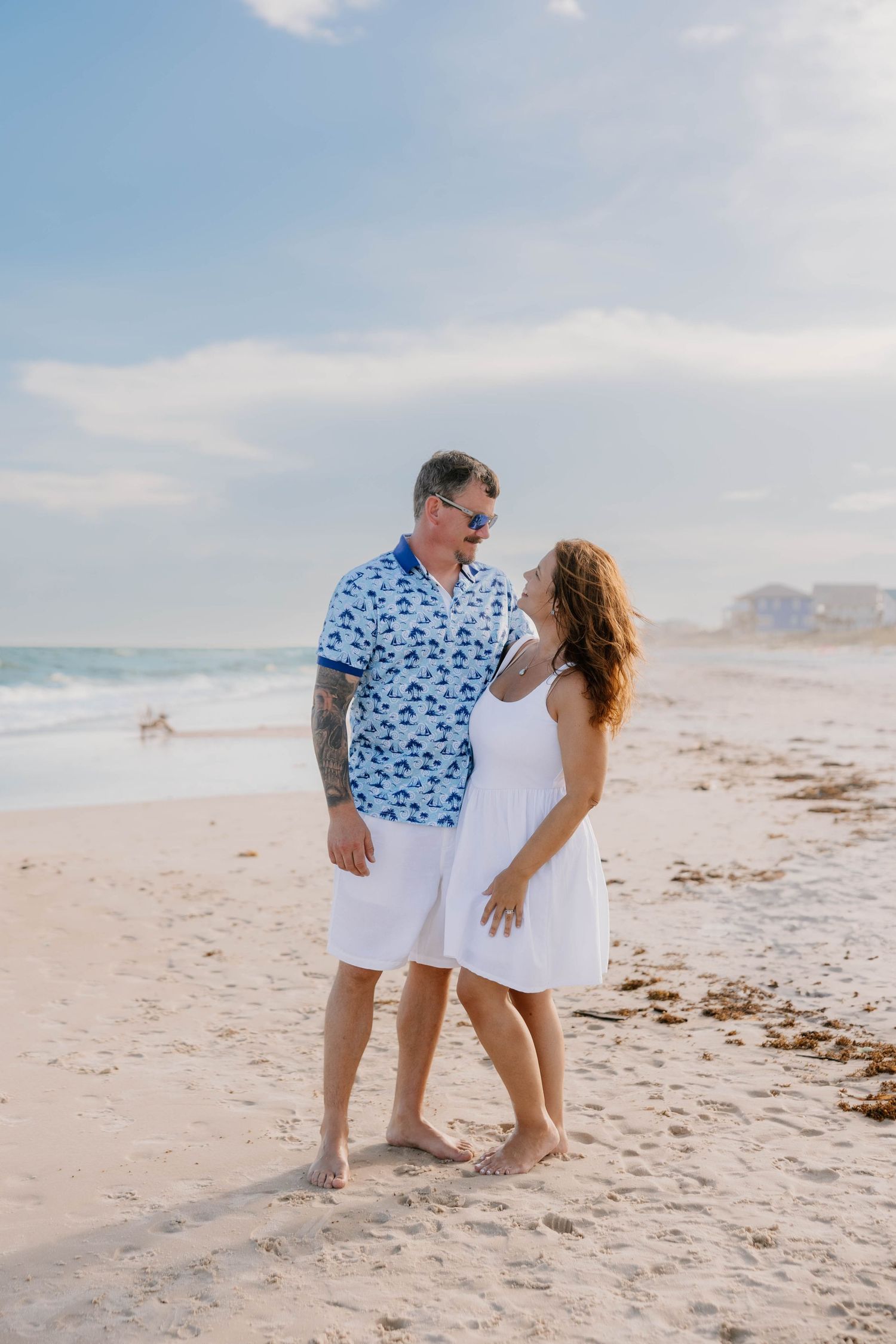 Couple in white and blue outfits sharing romantic moment on sandy beach.