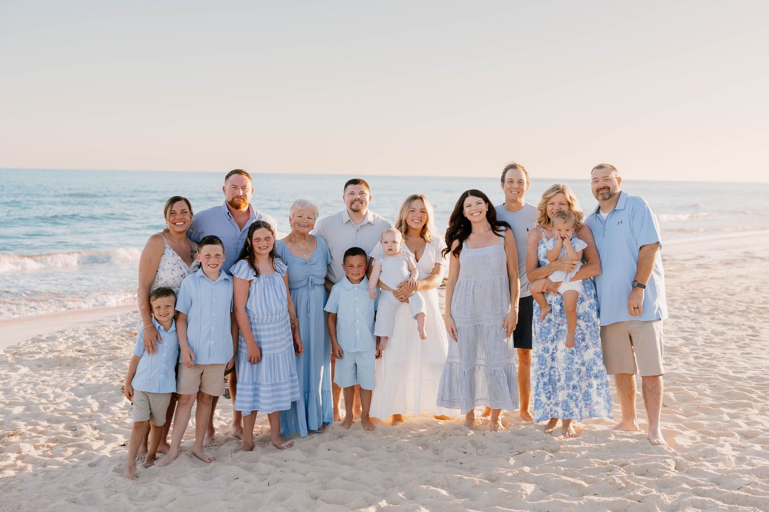 Large family group posing together on a sandy beach at sunset, wearing coordinated blue and white outfits.