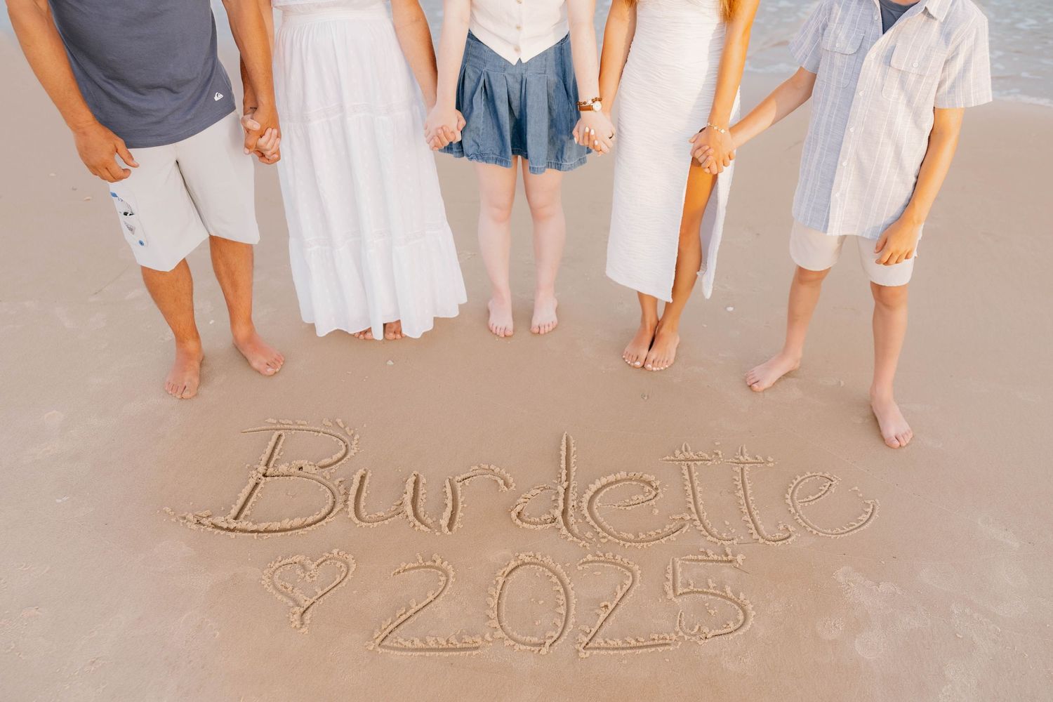 Family of five standing on beach with Burdette 2025 written in sand, holding hands at ocean shore.