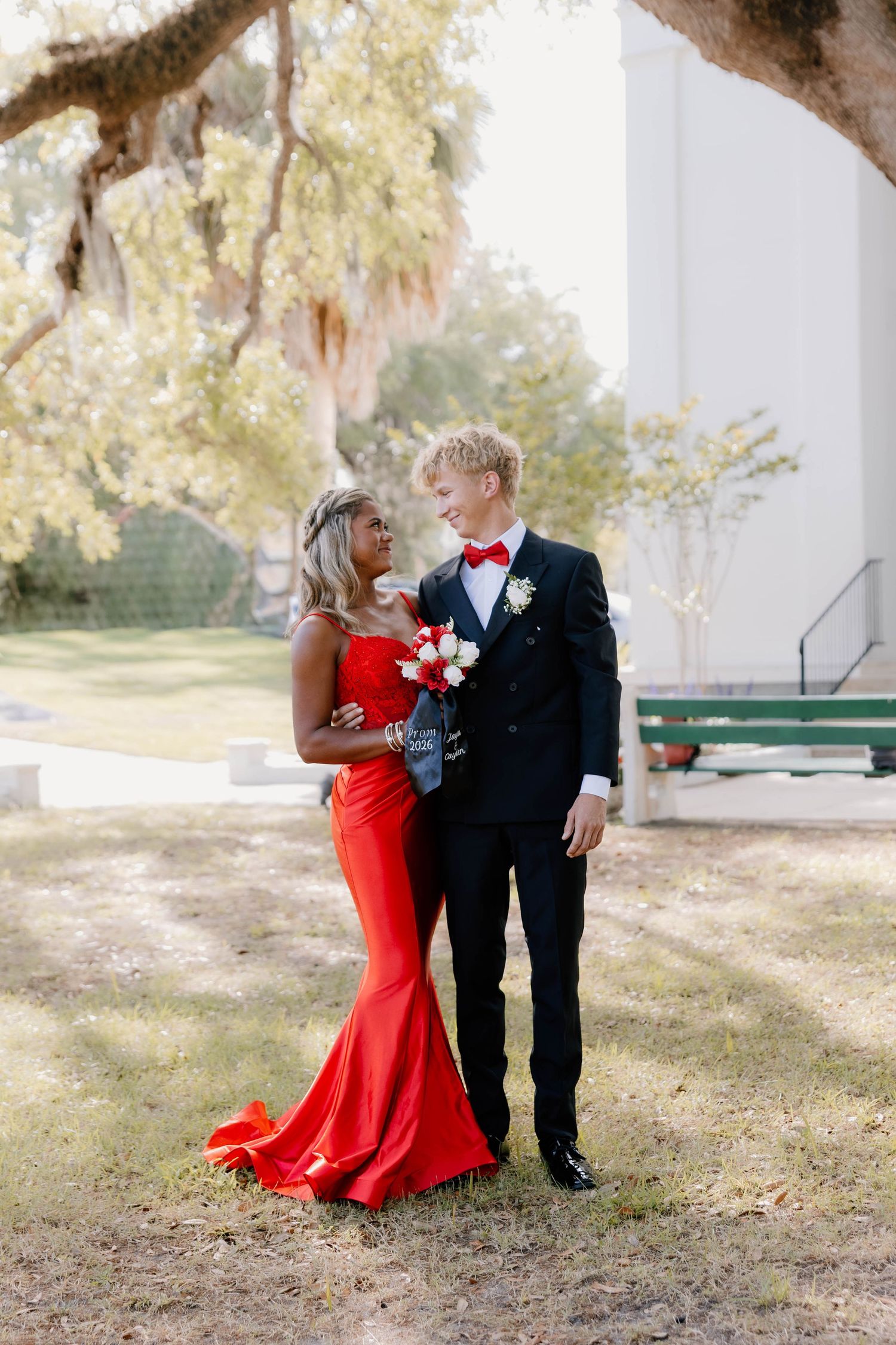 A couple in formal attire, woman in red gown holding bouquet, man in black tuxedo with red bow tie, posing outdoors.