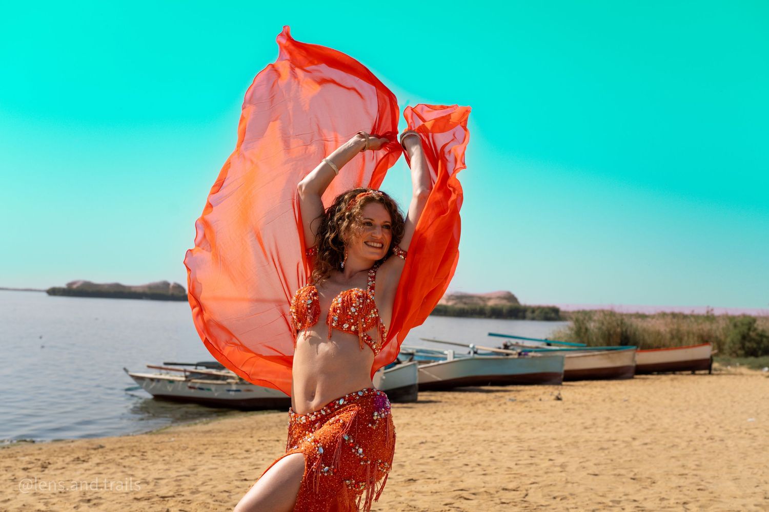 Belly dancer in orange costume waves red fabric on sandy beach with boats and blue sky behind her.