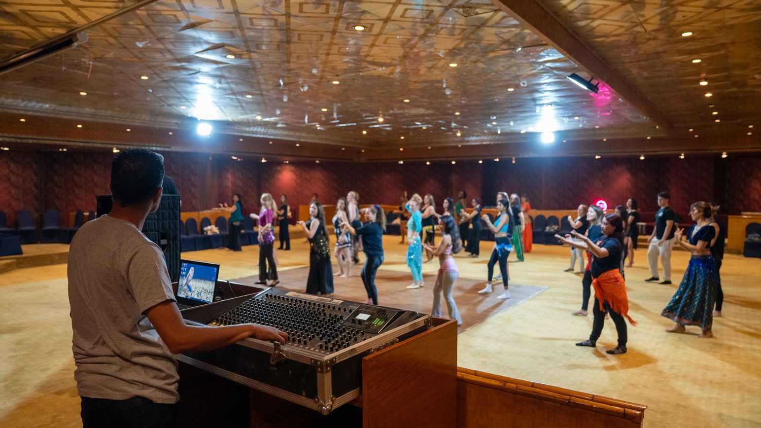 DJ operates sound mixing board while dancers rehearse in large ballroom during dance festival workshop.