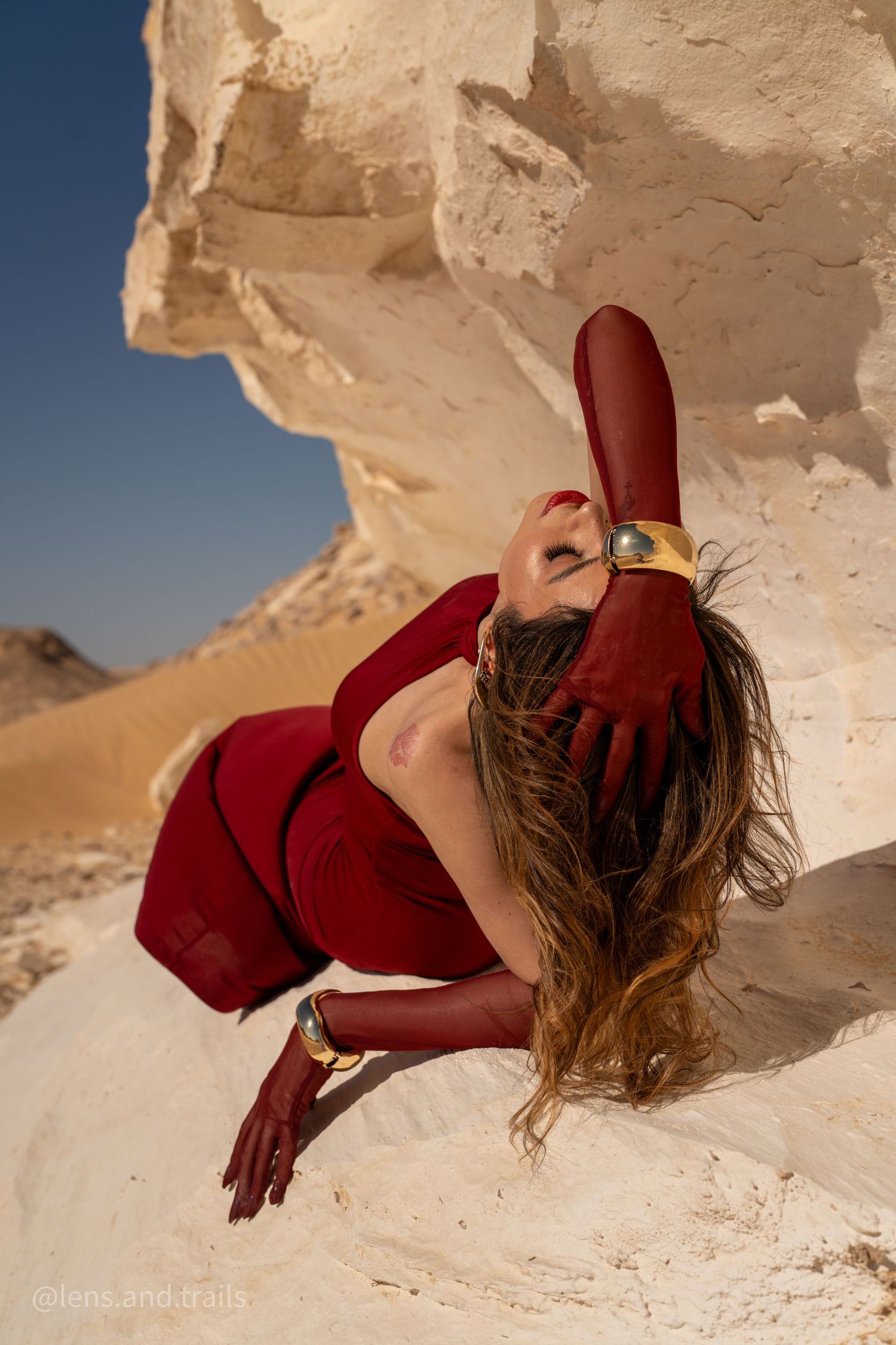 Model in red dress reclines on white desert rock with gloved hand raised upward.