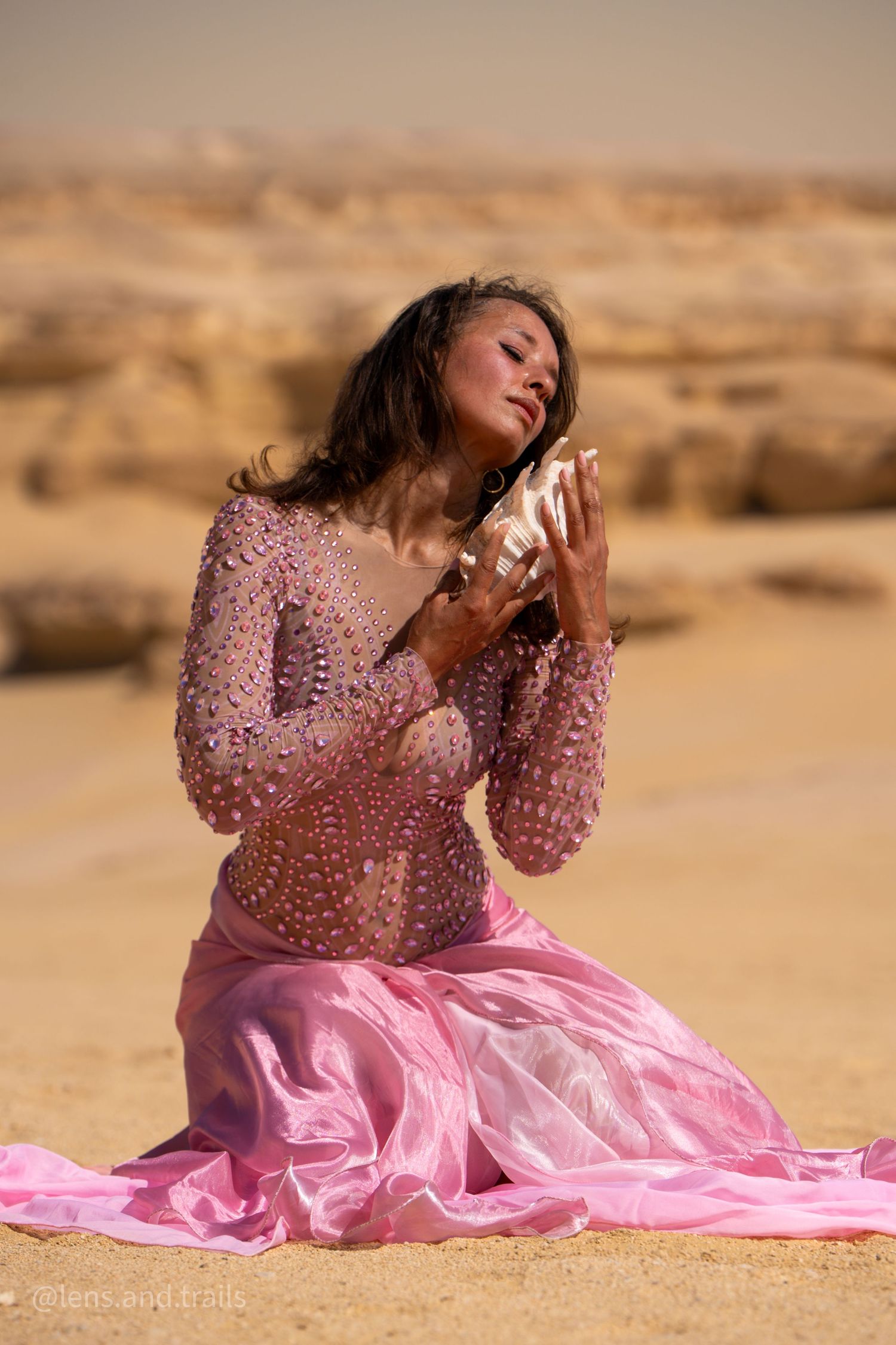 Woman in pink beaded gown kneeling on desert sand holding seashell with eyes closed peacefully.
