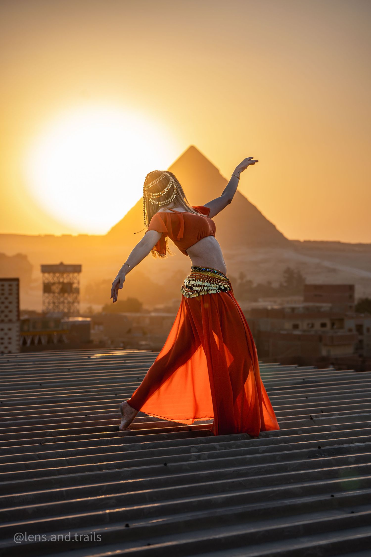 Belly dancer in red outfit with braided hair performing on rooftop with Egyptian pyramid at sunset.