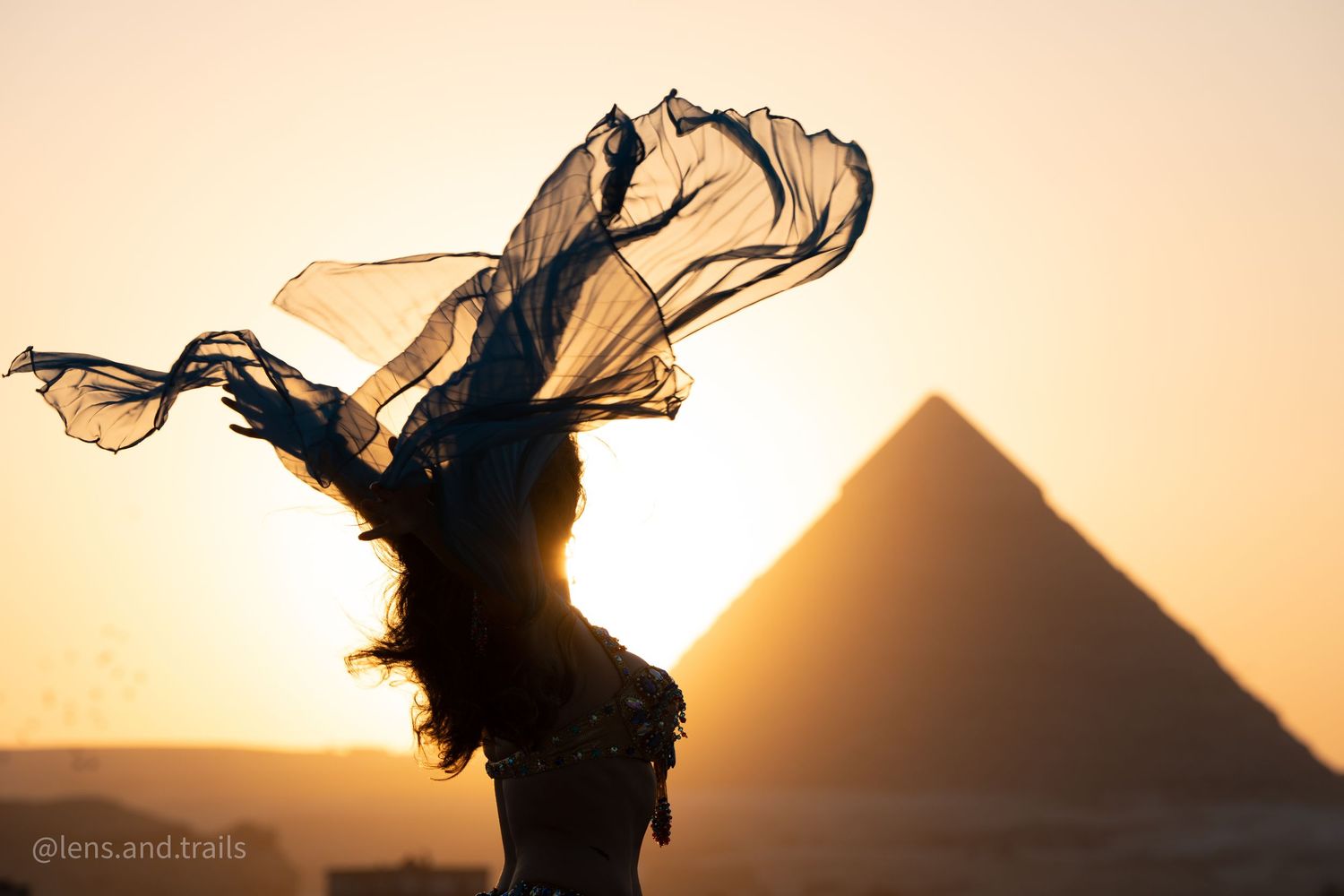 Silhouette of belly dancer with flowing veil against pyramid at golden sunset.