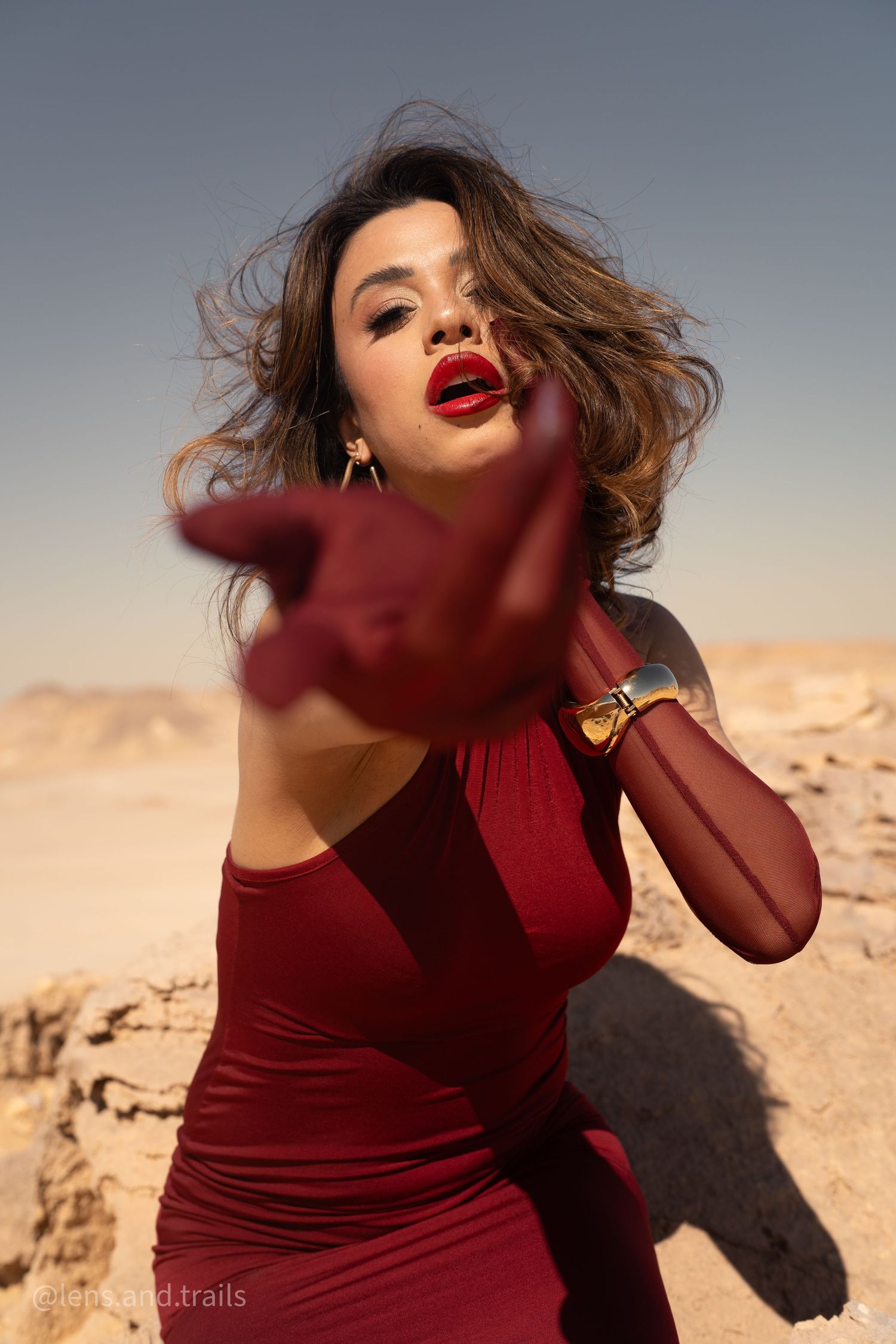 Close-up of woman in red dress with bold red lips reaching gloved hand toward camera in desert.