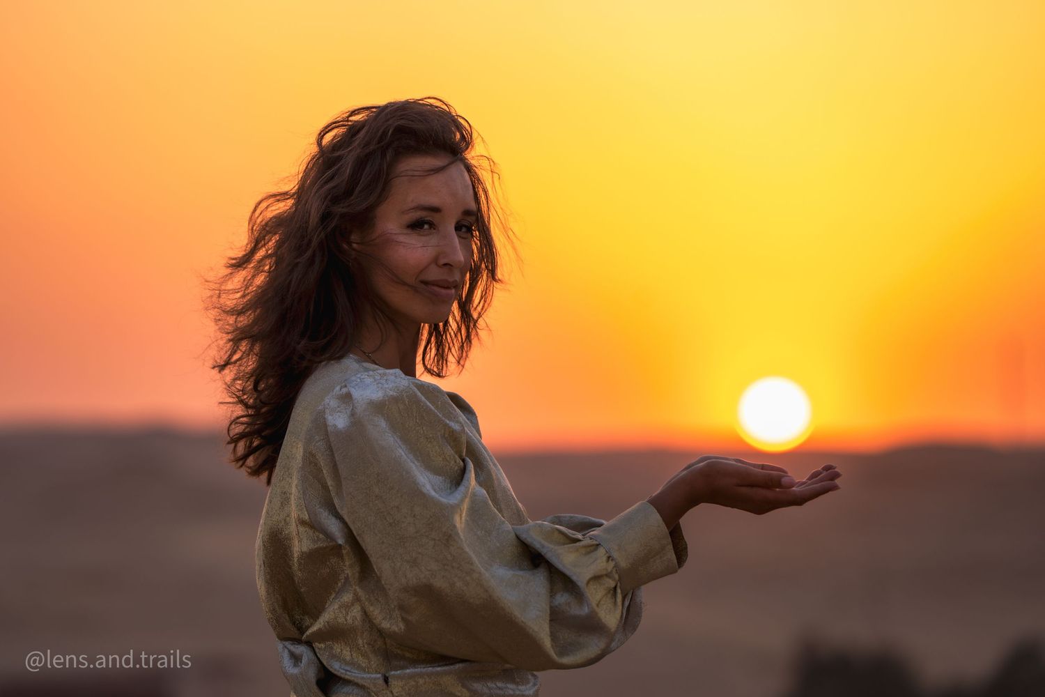 Smiling woman in gold outfit appears to hold the sun in her palm at desert sunset.