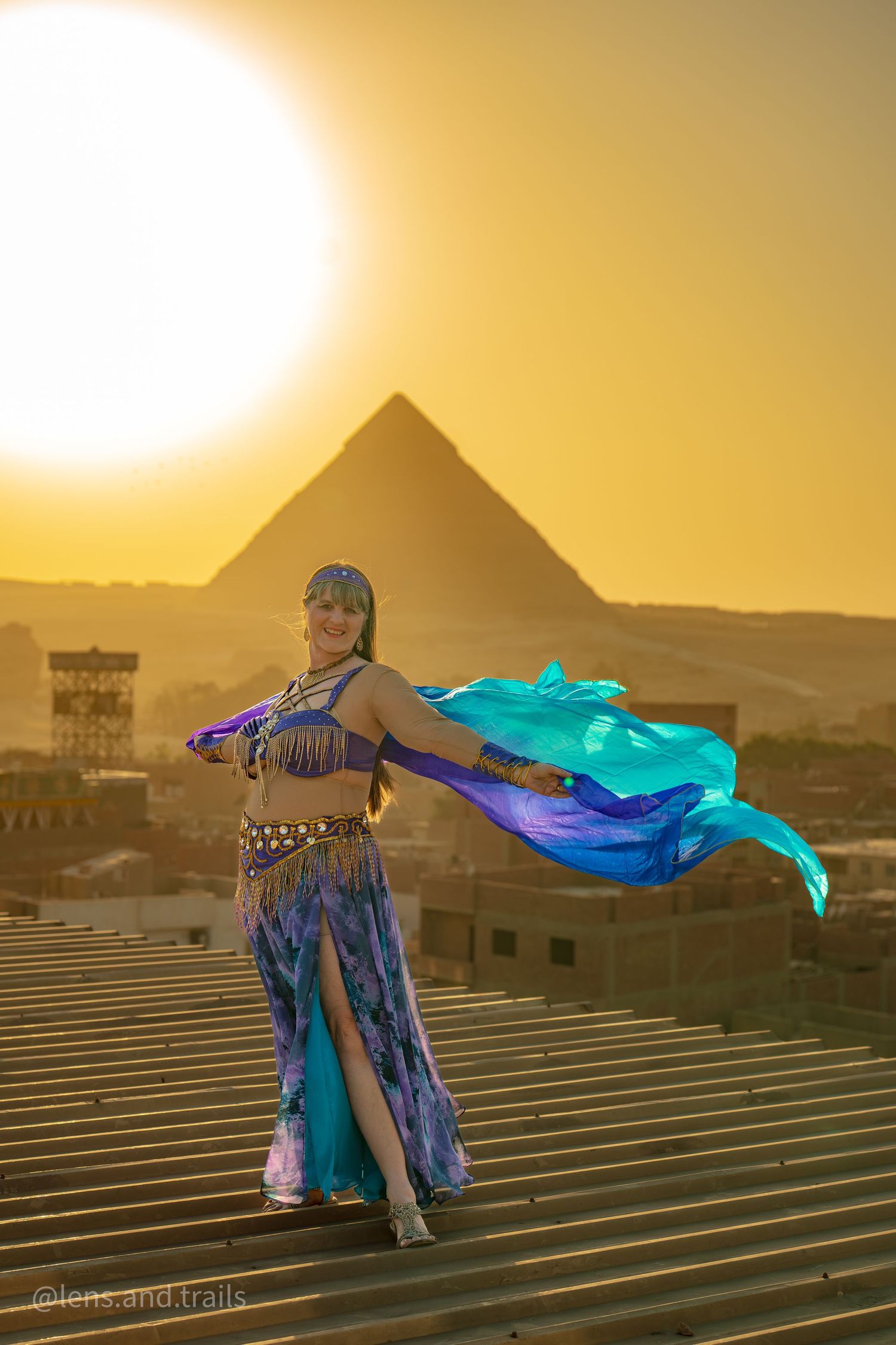 Belly dancer in blue and purple costume with silk veils performing near Egyptian pyramids at sunset.