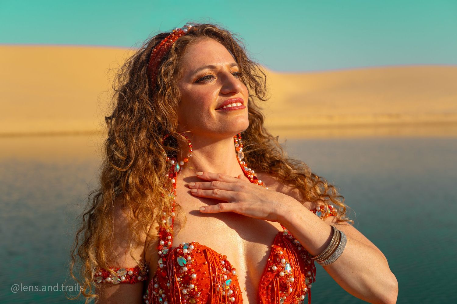Close-up of smiling belly dancer in red beaded costume with curly hair near desert oasis water.