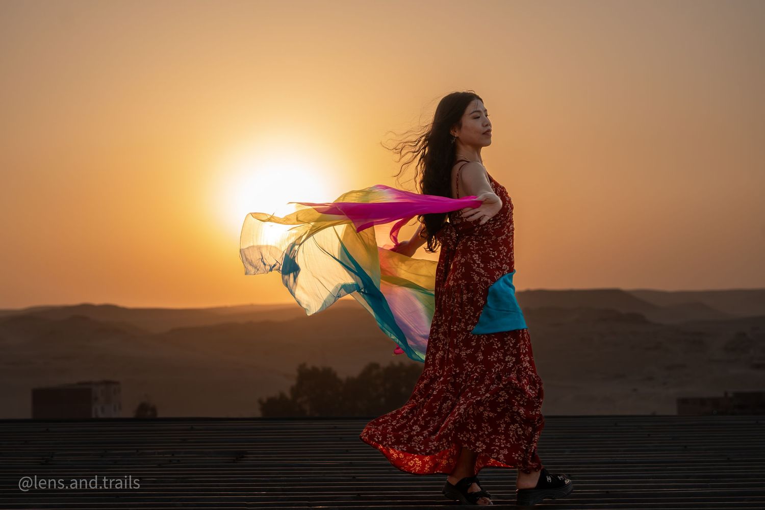 Woman in red floral dress twirls colorful scarf at desert sunset.