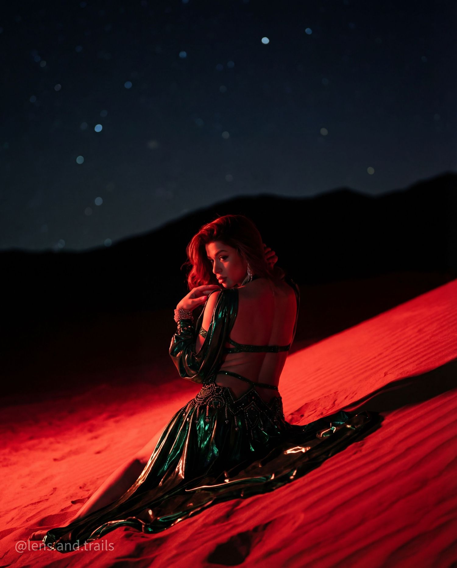 Woman in green sequined costume sits on red-lit desert sand dunes under a starry sky.