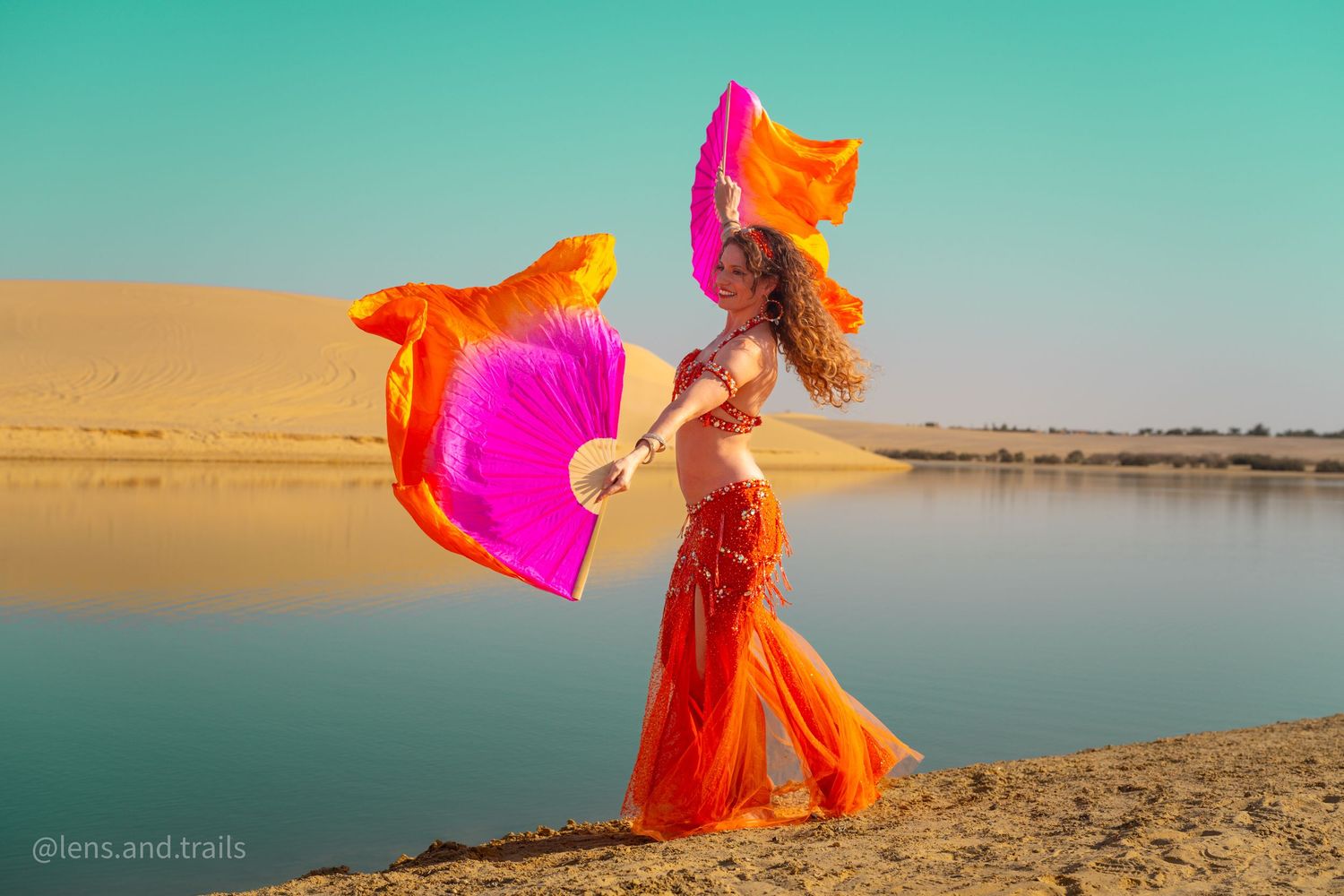 Belly dancer in orange outfit waves pink and orange fans beside a desert oasis with golden dunes.
