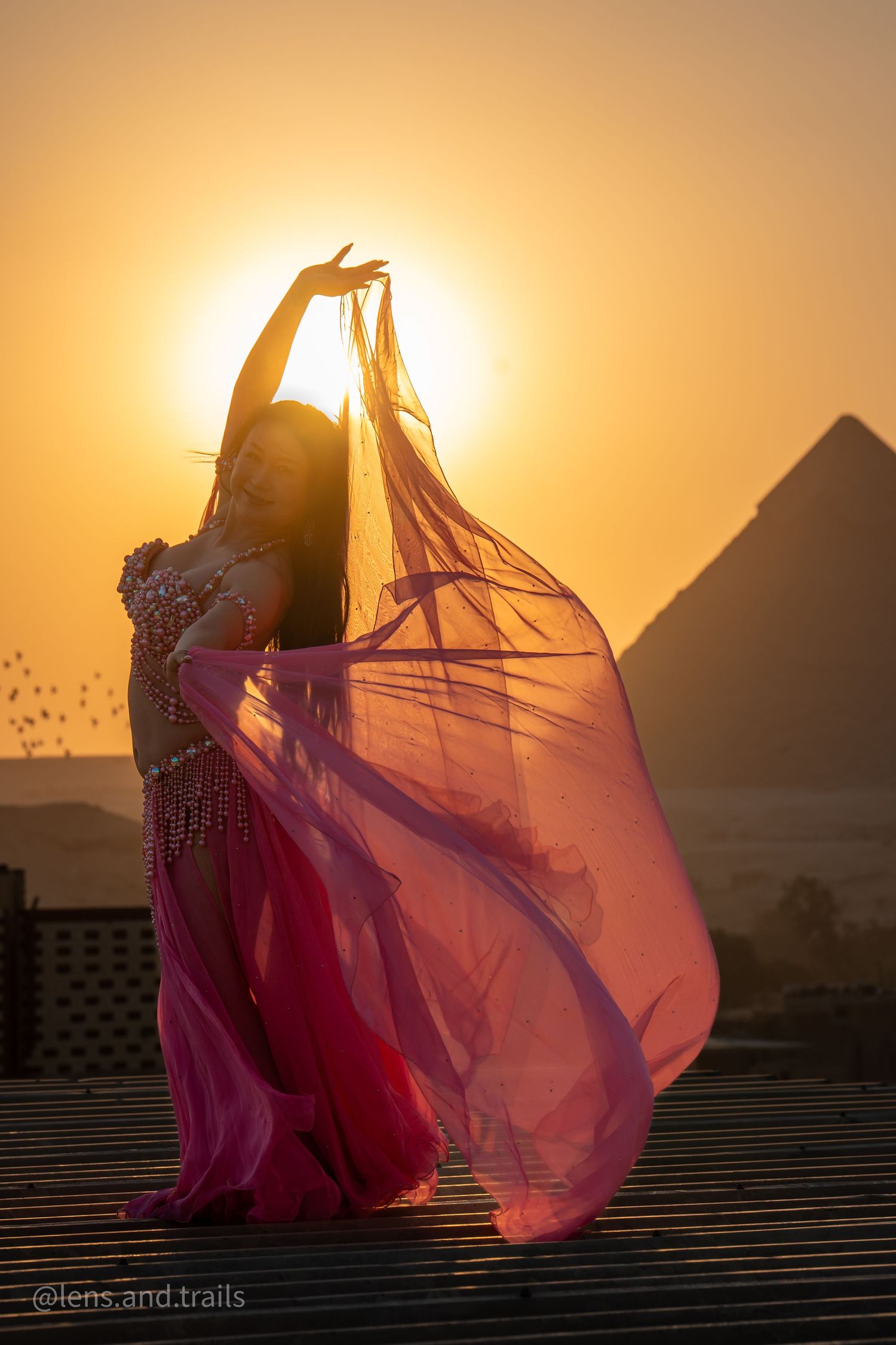 Belly dancer in pink costume twirling flowing veil before the Great Pyramid during golden sunset.