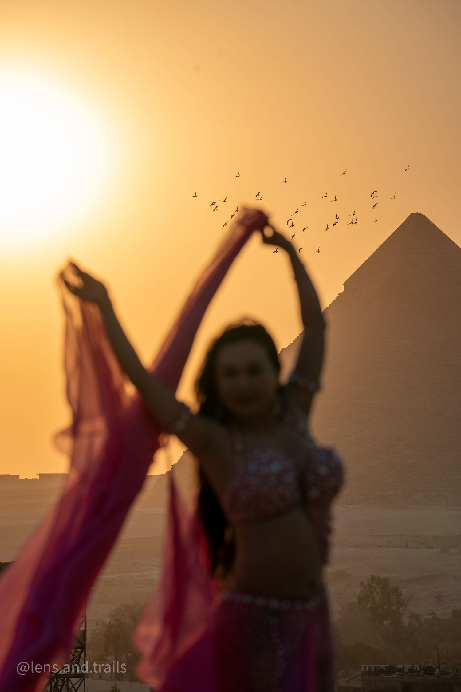 Dancer in pink belly dance costume raises veil before pyramid at sunset.