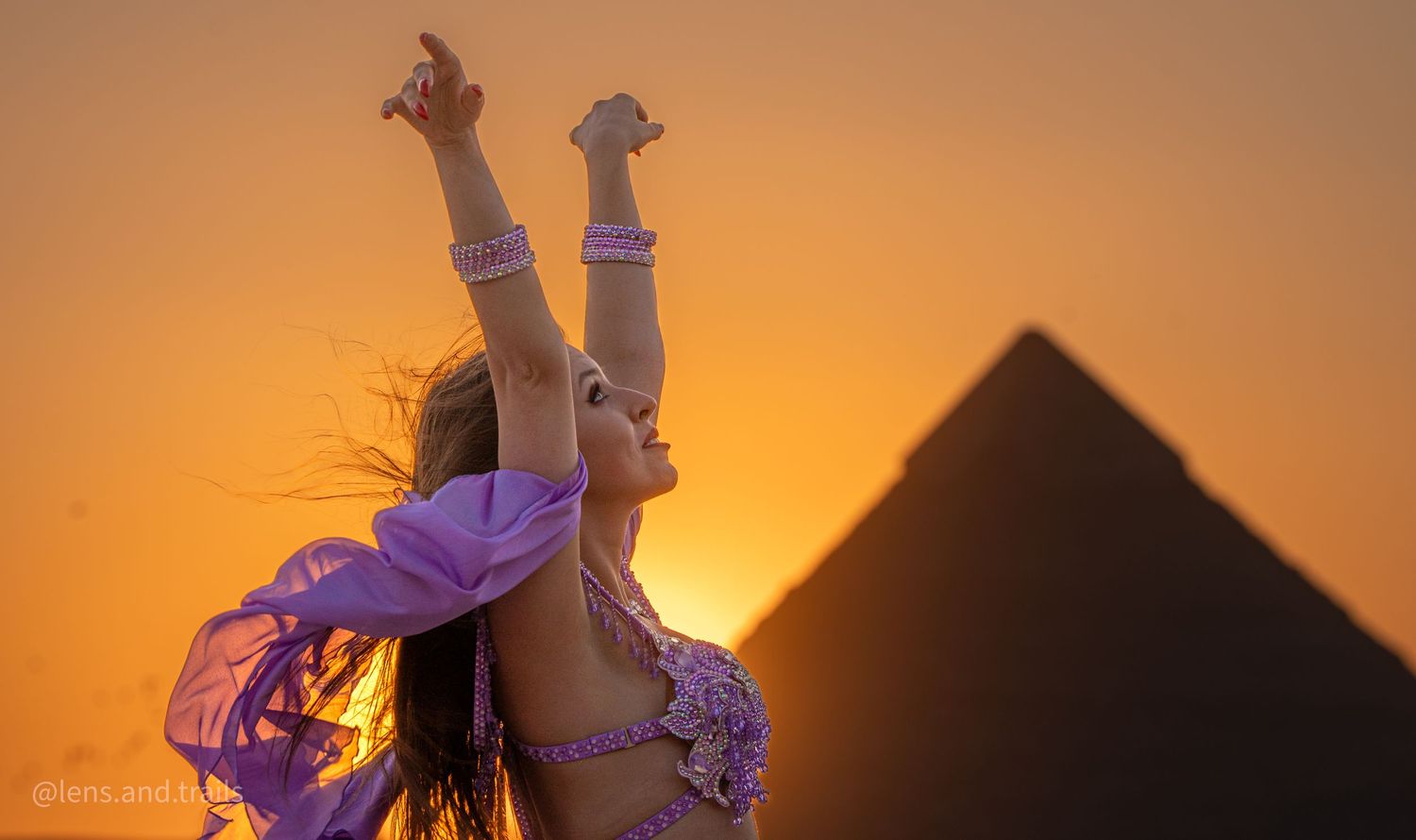 Belly dancer in purple costume with arms raised against Egyptian pyramid at sunset.