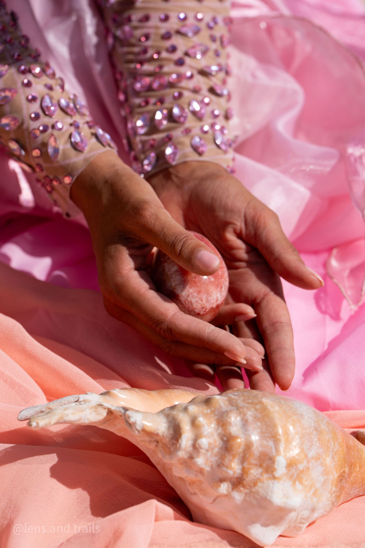 Close-up of hands holding pink rose quartz crystal beside a large seashell on pink fabric.