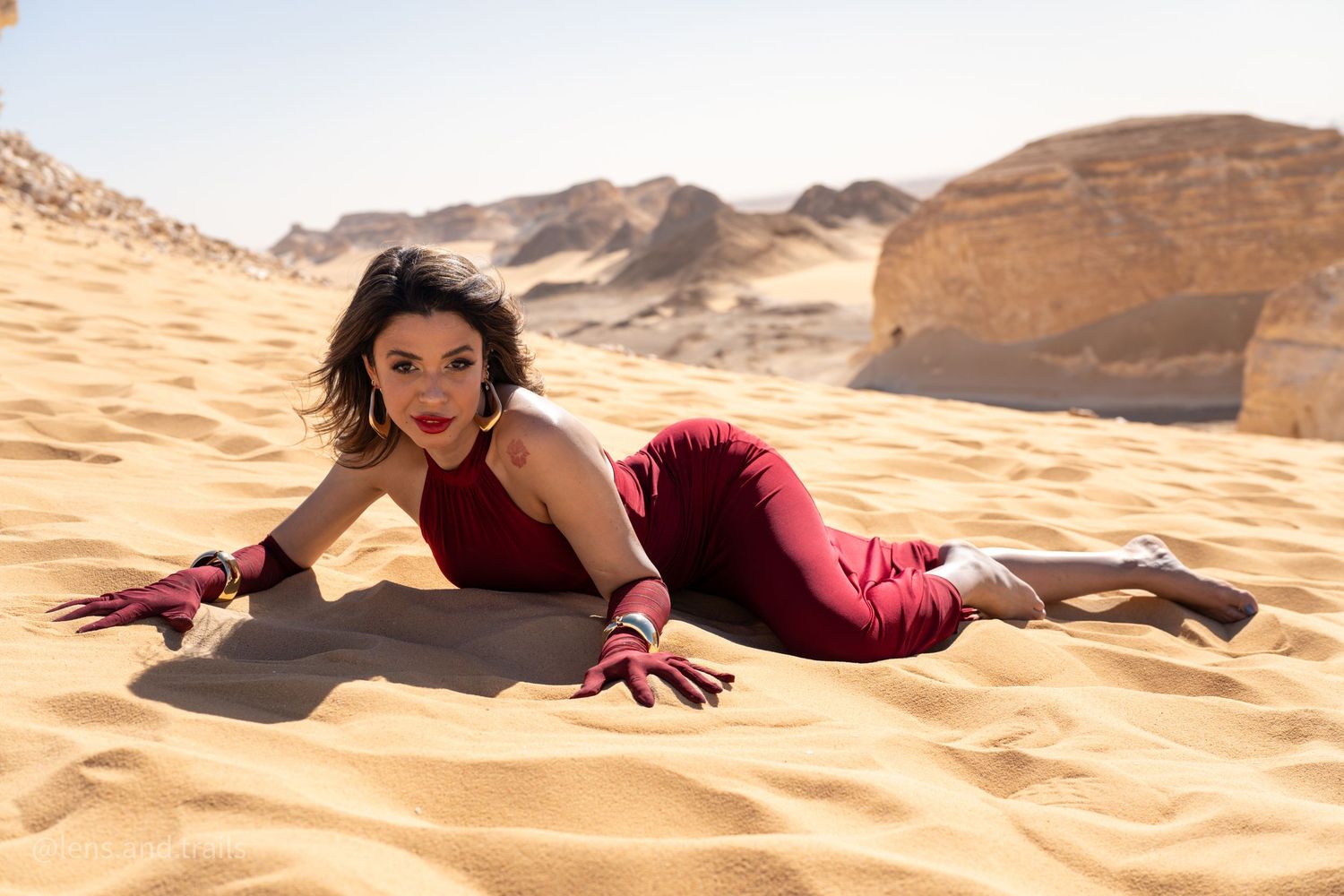 Woman in red outfit lies on desert sand with arms outstretched in a dramatic pose.