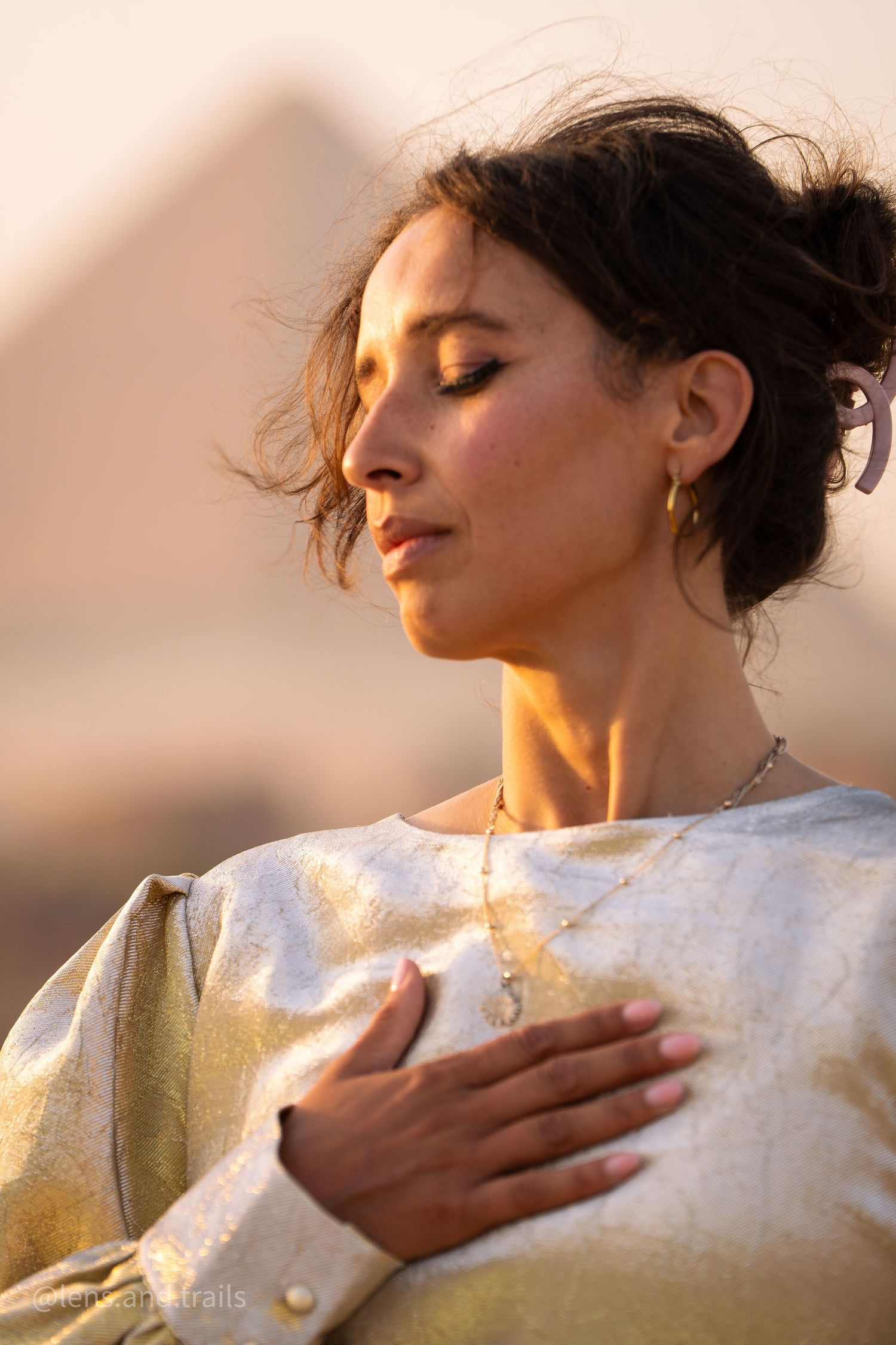 Woman in gold dress with eyes closed, hand on chest, pyramids softly blurred behind.