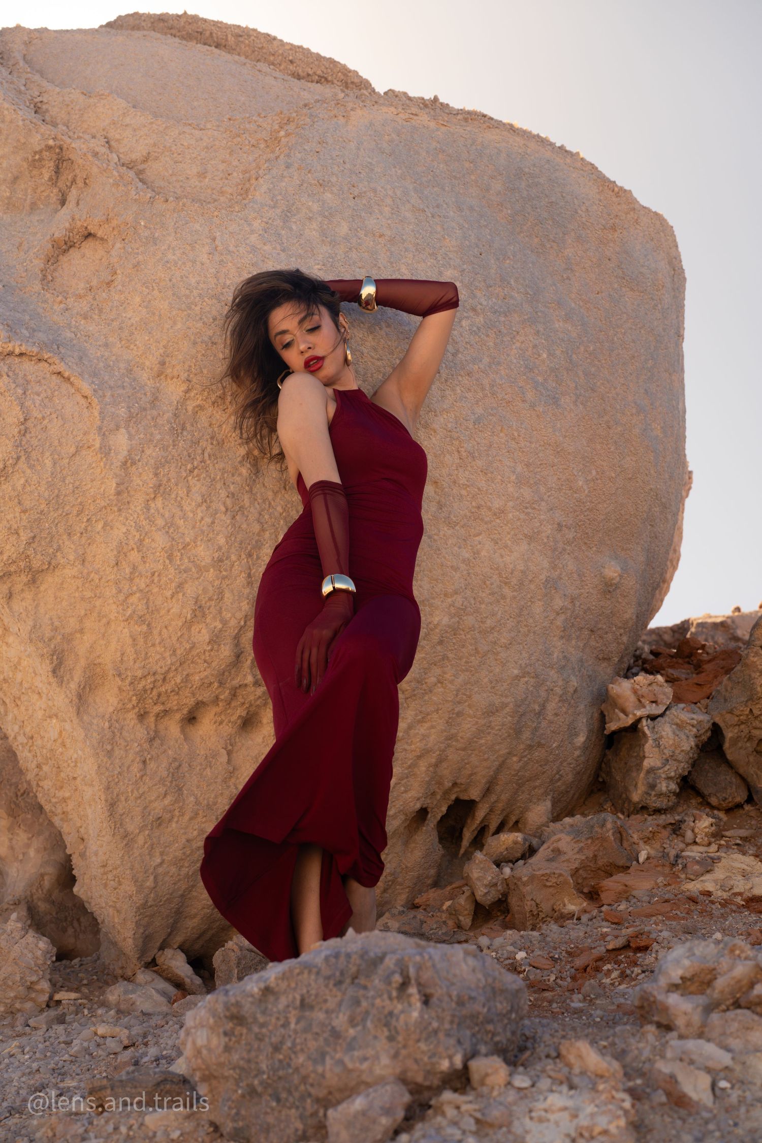 Model in crimson dress leans against large boulder in arid rocky desert setting.