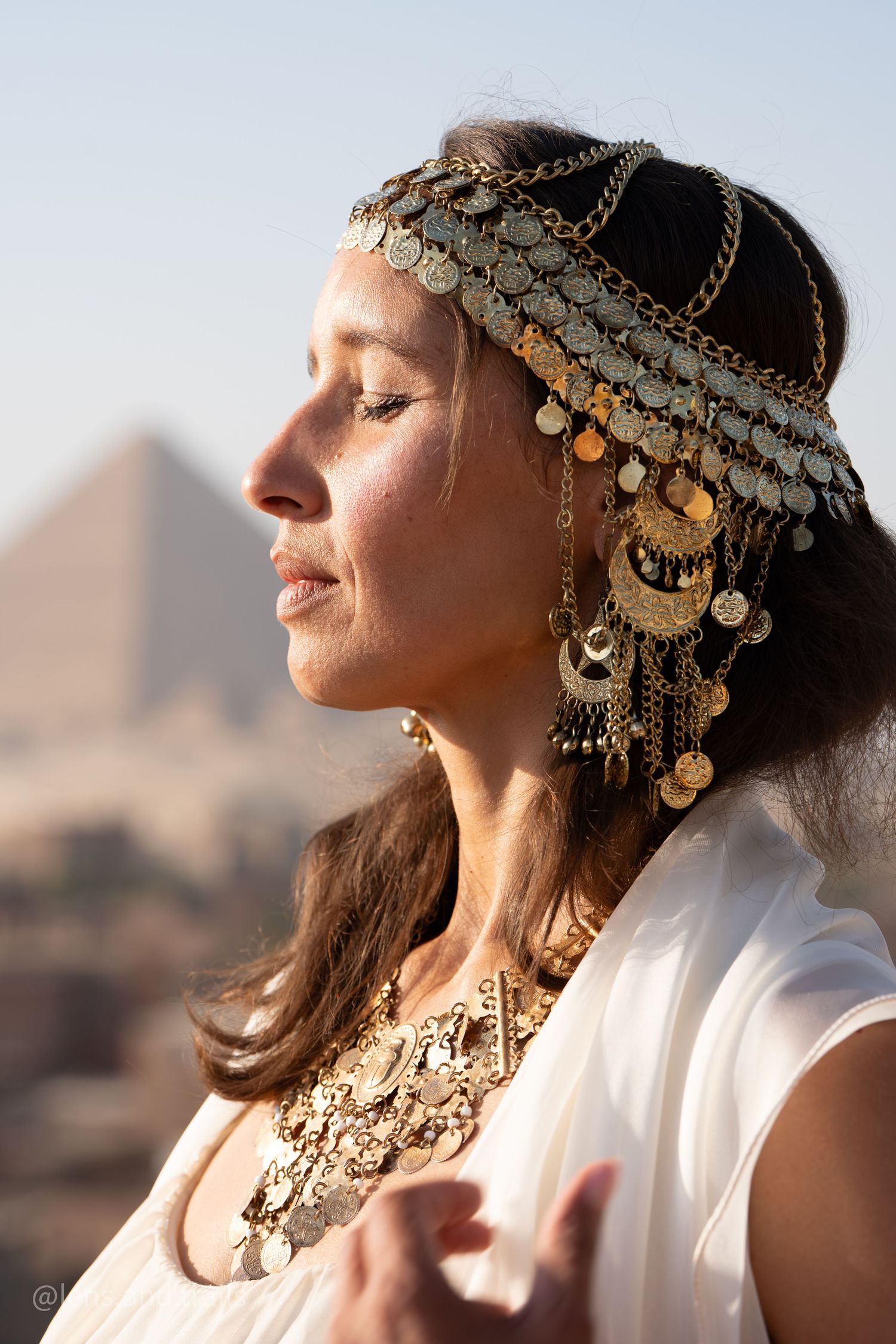 Woman in golden coin headpiece and necklace meditates before Egyptian pyramids.