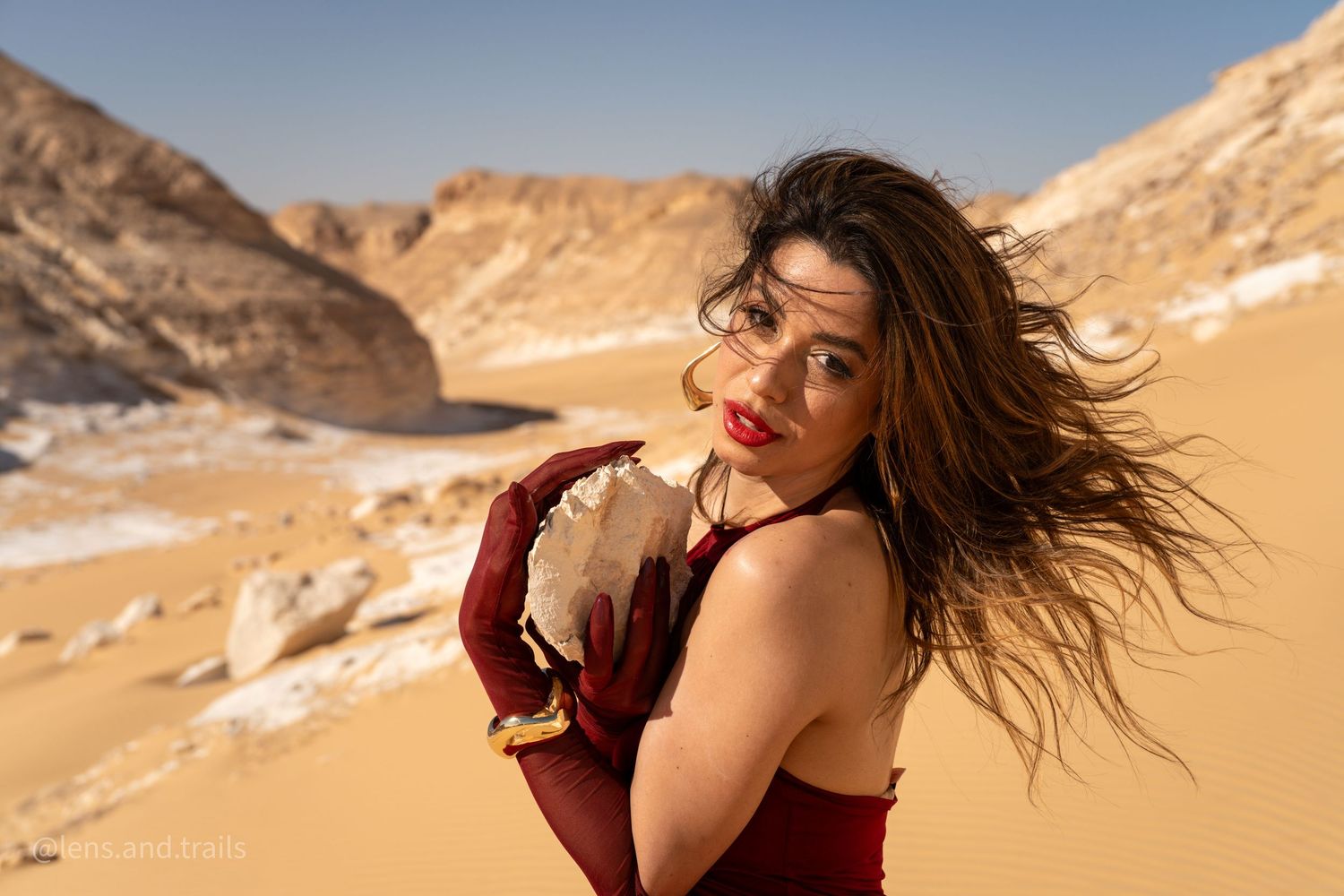Close-up of brunette woman in red dress holding desert rock with wind-blown hair.