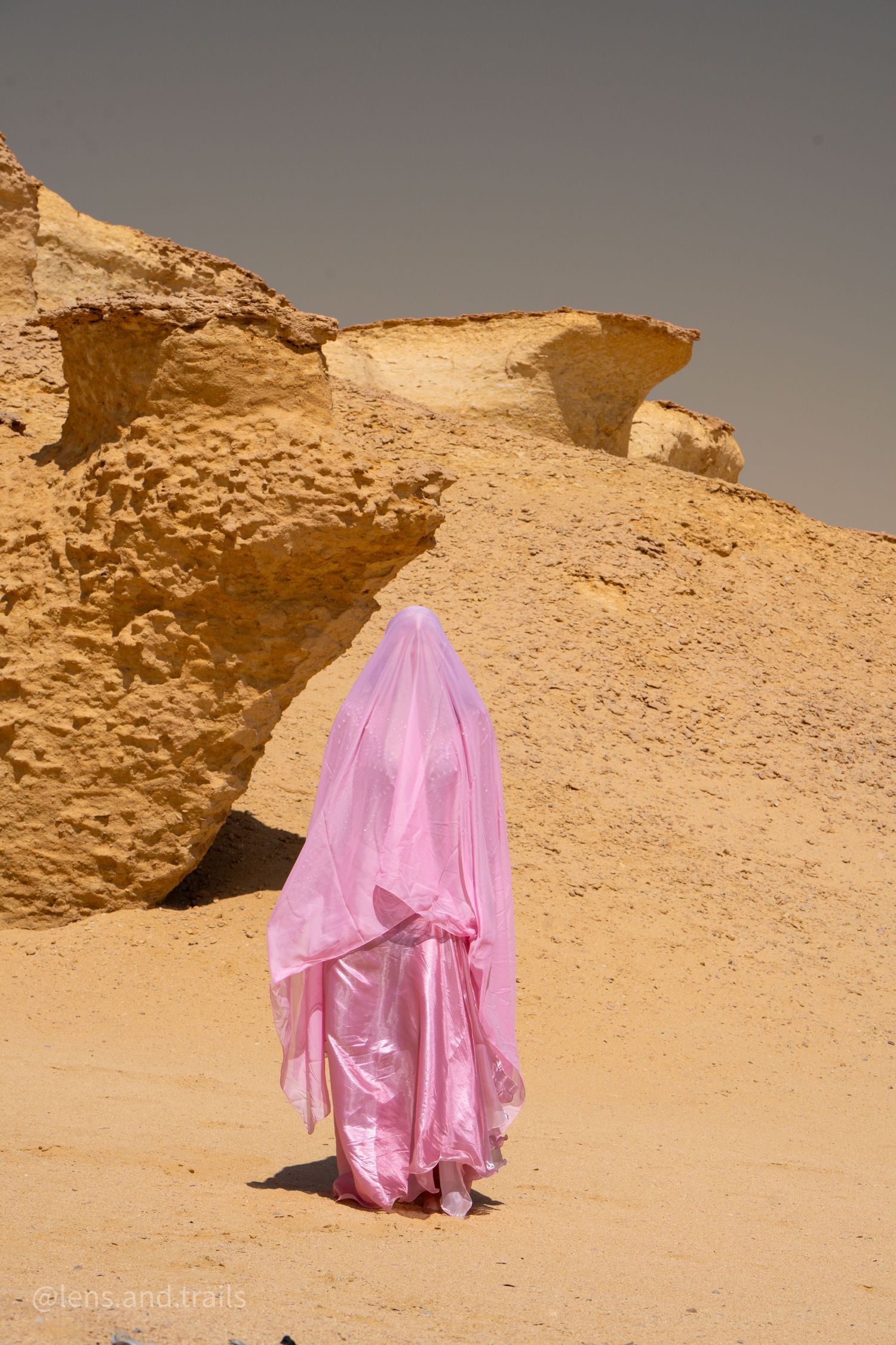 Figure draped entirely in flowing pink fabric walks through golden sandy desert rock formations.