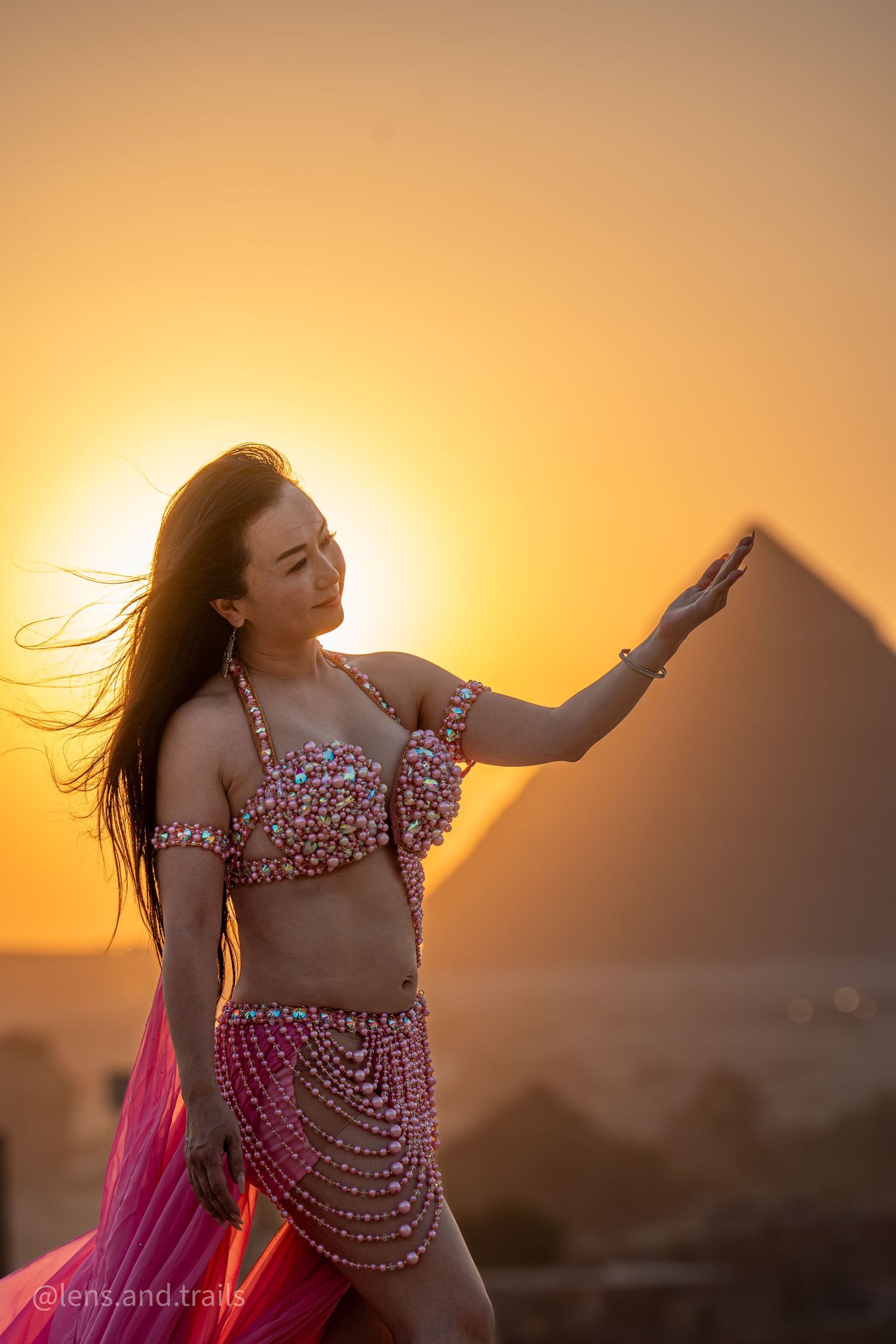 Belly dancer in ornate pink beaded costume posing gracefully before Egyptian pyramids at golden sunset.